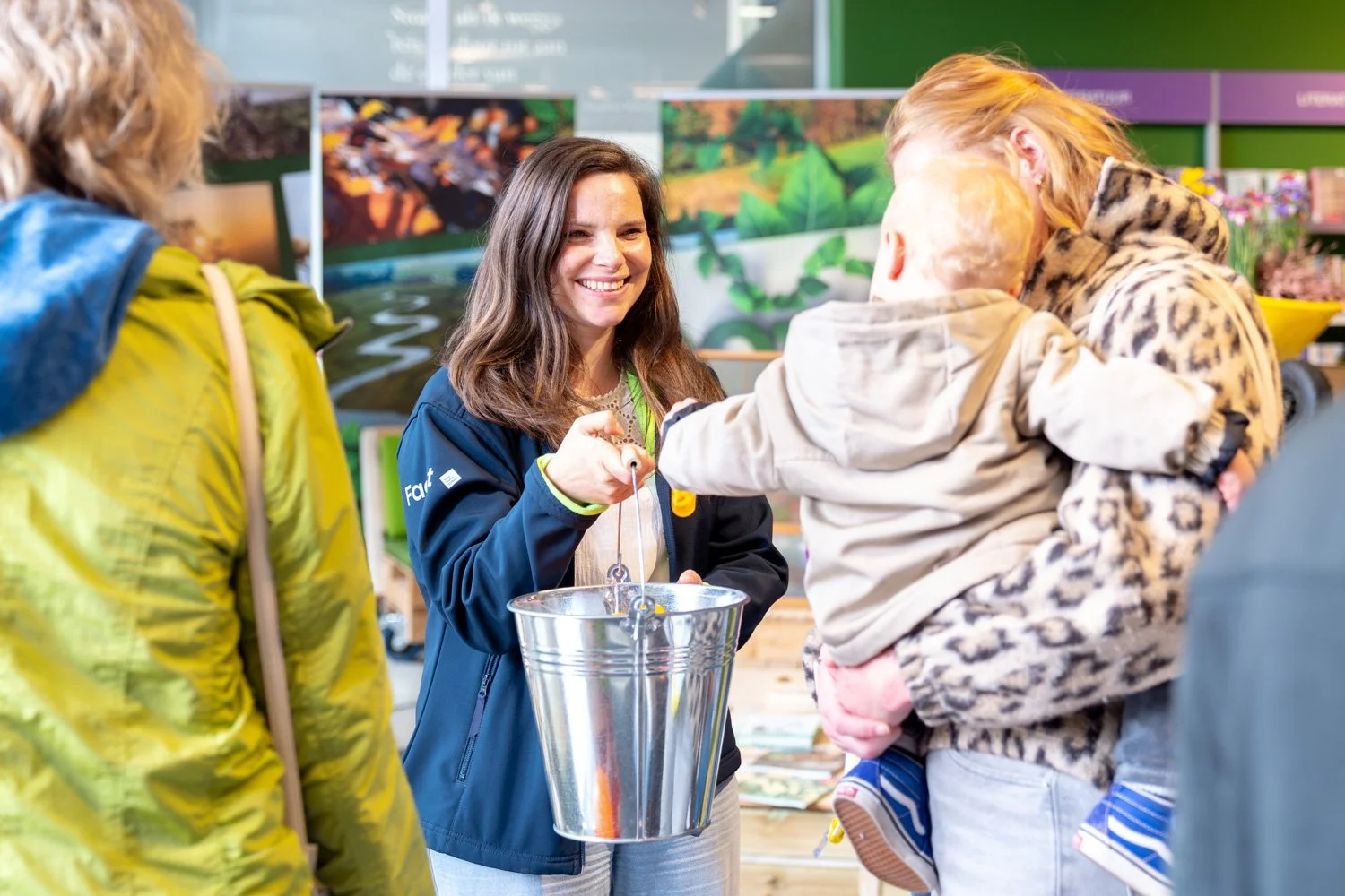 Vrouw die met kinderen in een winkel of markt staat, lachend en een metalen emmer vasthoudend.