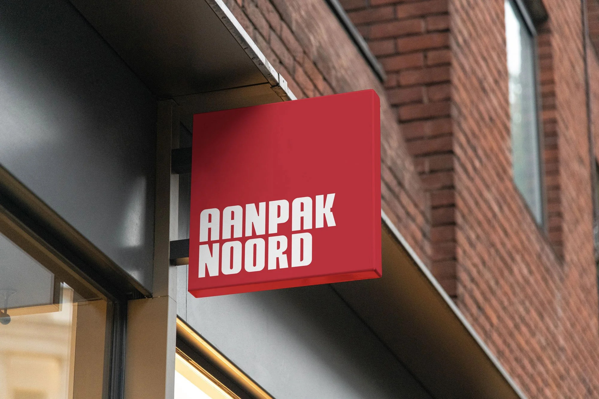 Groen en rood uithangbord met witte tekst 'Aanpak Noord' hangend aan een muur van een gebouw met bakstenen gevel.