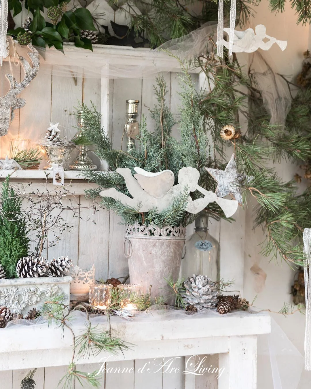 Juldekoration med grønne grene, pinecones, keramikfigurer af duer, stjerner og små lys, placeret på en hvid træbord med en hvid træbaggrund.