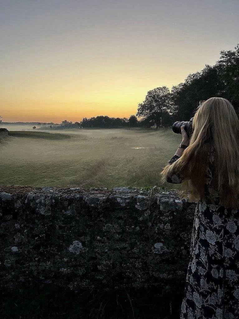 Kvinde fotograferer solnedgang over et tåget landskab med træer og mark.