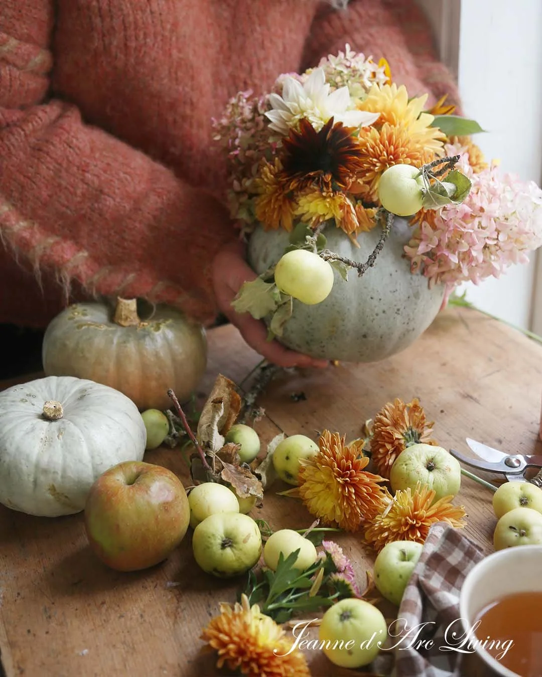 En person i en pink sweater holder en grå blomsterpotte med orange, hvid, gul og pink blomster samt små grønne æbler. Flere grønne æbler, hvide og grå pumpkins samt blomster er spredt ud over en træbord.