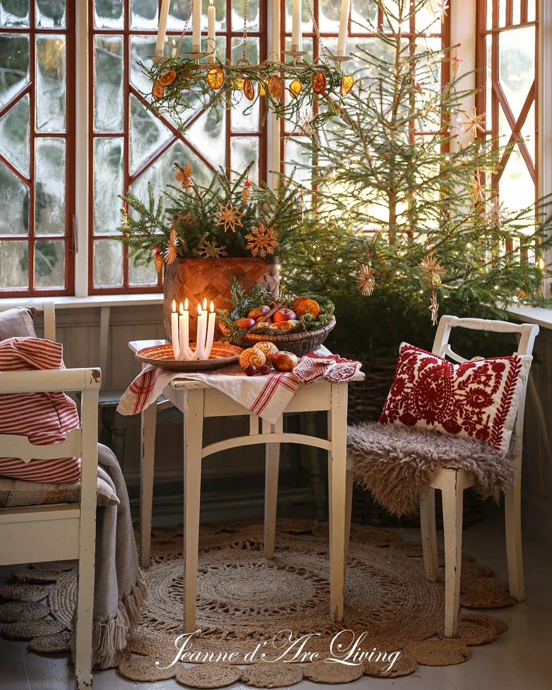 Juletræ med pynt i et værelse med store vinduer. En borddekoration med stearinlys, juledekorationer, frugt og snefnug. Der er julehjerter og tekstiler i rødt og hvidt.