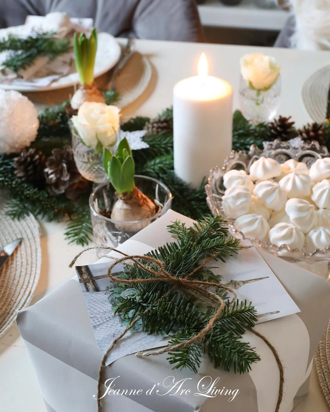 Juledekoration med grønne grene, lys, hvide blomster, små kogler og kage med marengs på et pyntet bord.