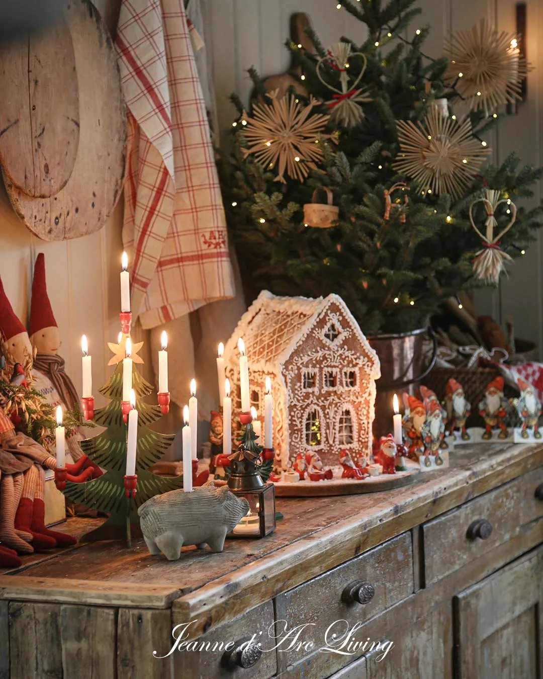 Juledekorationer på en træoverflade med en adventskrans med lys, en uldhytte pyntet med icing, små juleunger, julefigurer og en juletræ med hjerteformede og stjerneformede dekorationer, samt nogle julemandfigurer og en lille gris.