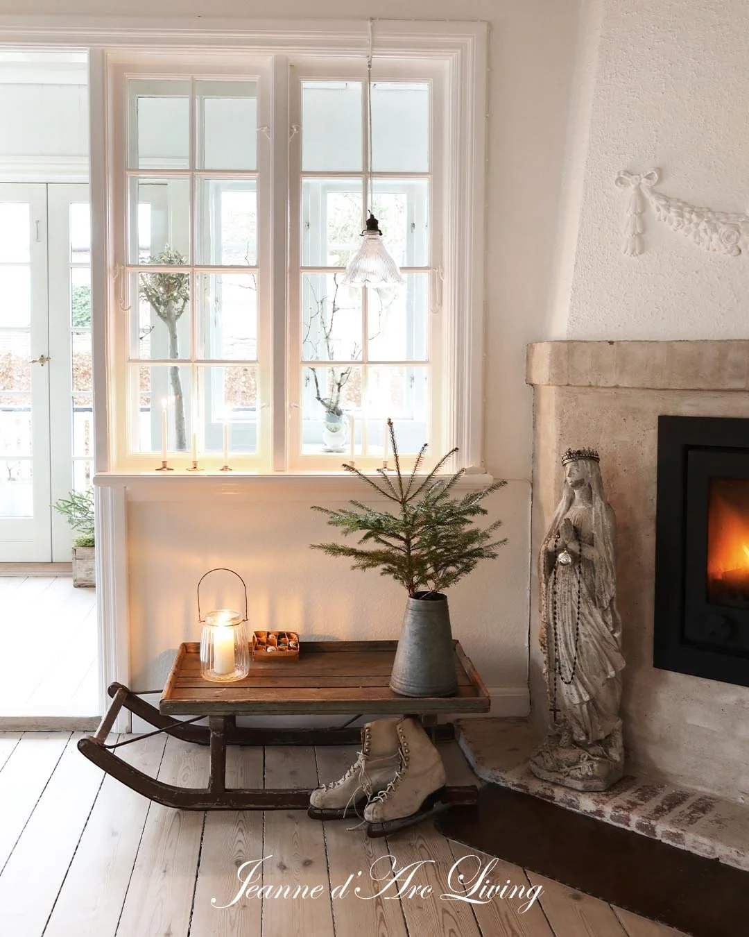 Indendørs stuehjørne med et lille grantræ i en grå vase på et træskohjulsbord foran en stor vindue, en stearinlygte, en tændt brændeovn, en juledekoration, og et par gamle skøjter under bordet. Der er også en figur ved siden af ovnen.