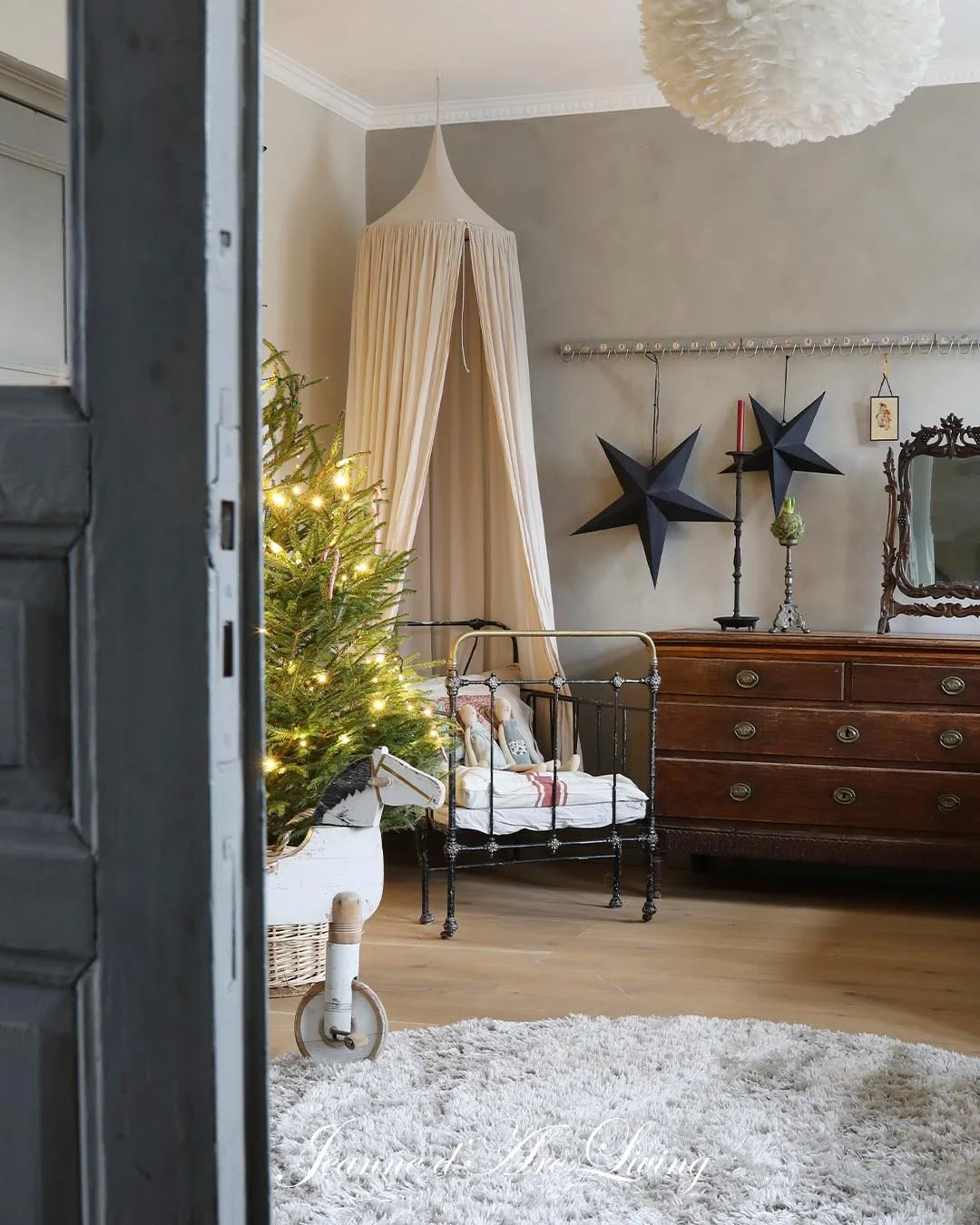 Indretning af et hyggeligt stue med juletræ pyntet med lys, en gammel seng med dukker, en trækommode med to sorte stjernedekorationer, en spejl og en beige gardin, set gennem en døråbning.