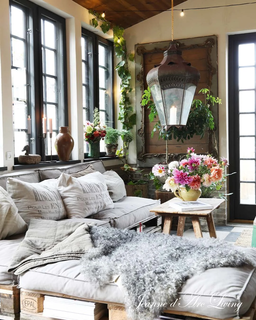 Indendørs stue med store vinduer, pufskud, blomsterarrangementer, en stor lysekrone, en vægdekor, og et sofabord. Der er grønne planter og levende lys. Rummet har en hyggelig og rustik stemning.