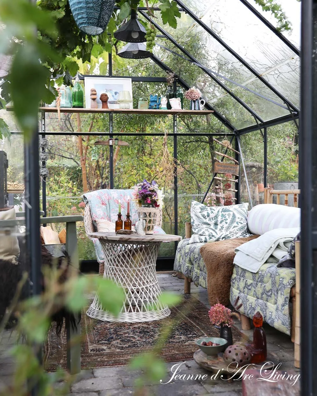 Indendørs af en hyggelig, botanisk-have-værelset med sofa, stol, blomster og dekorative genstande, set gennem en glasindhegning.