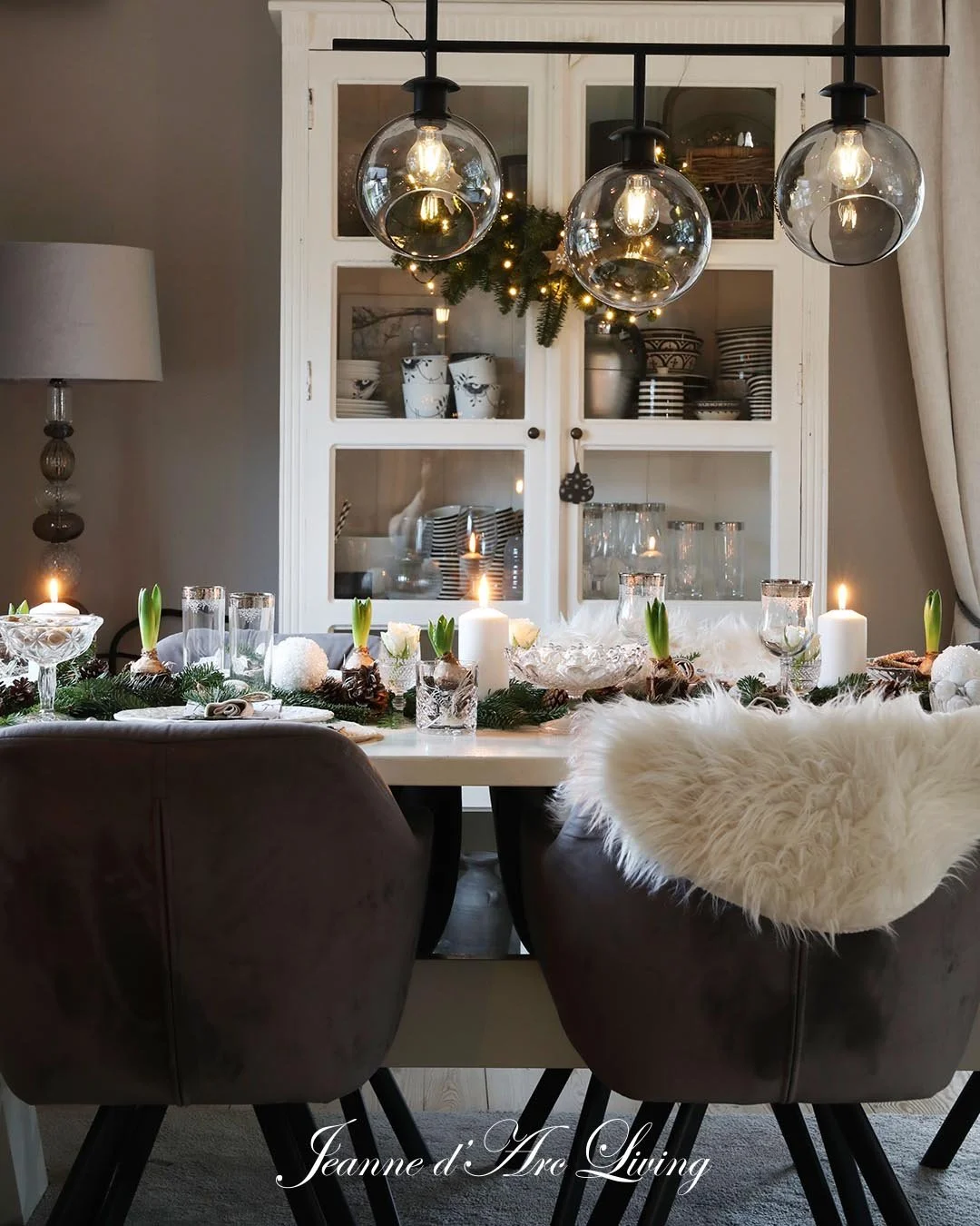 Juledekoration på spisebord med lys, blomster, gran, og stearinlys, over en hvid cabinet med tekopper og glas