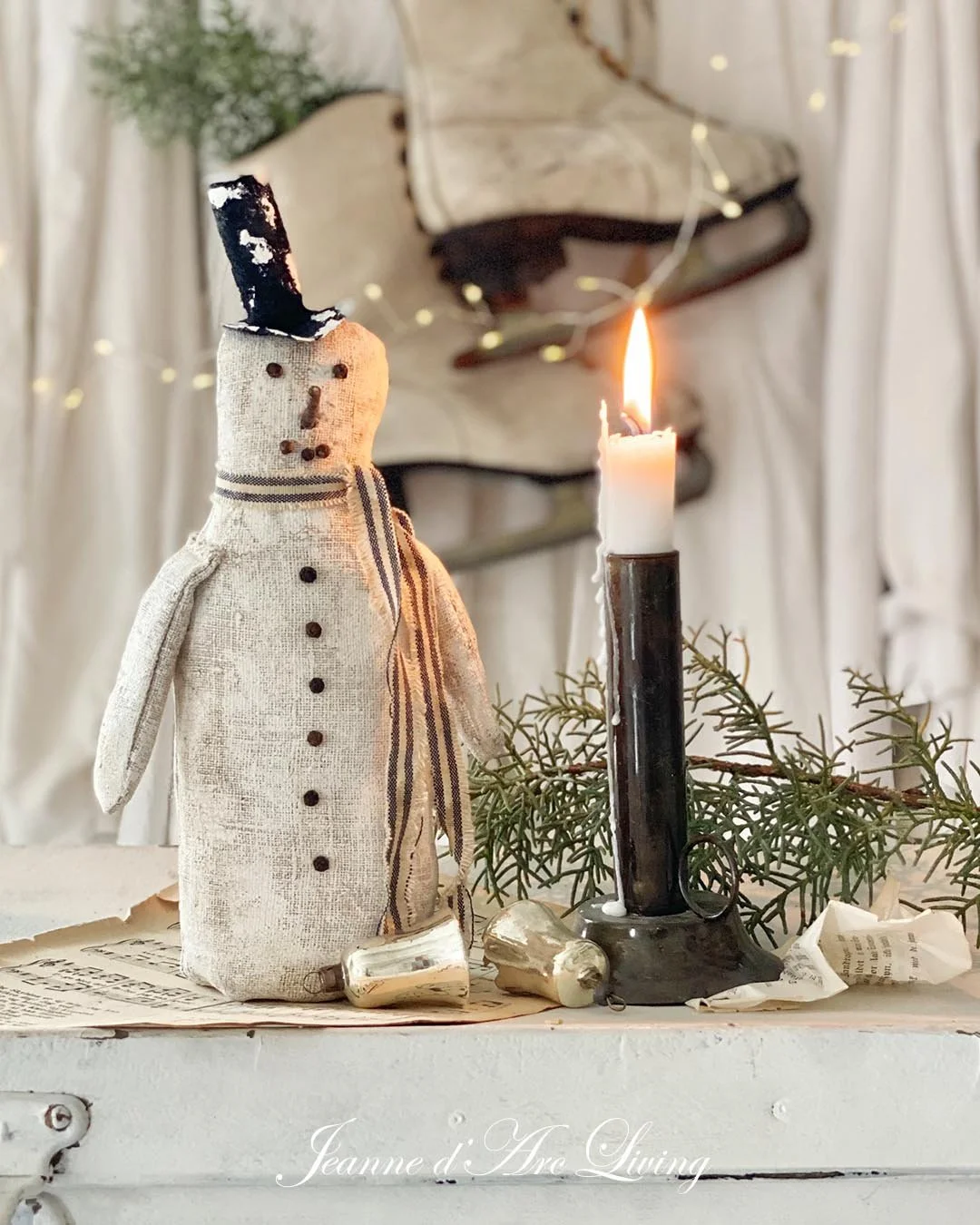 En juledekoration med en hjemmelavet snemand lavet af tøj, en sort stearinlys og grønne grene. I baggrunden er der en væg med julelys og en træskulptur.