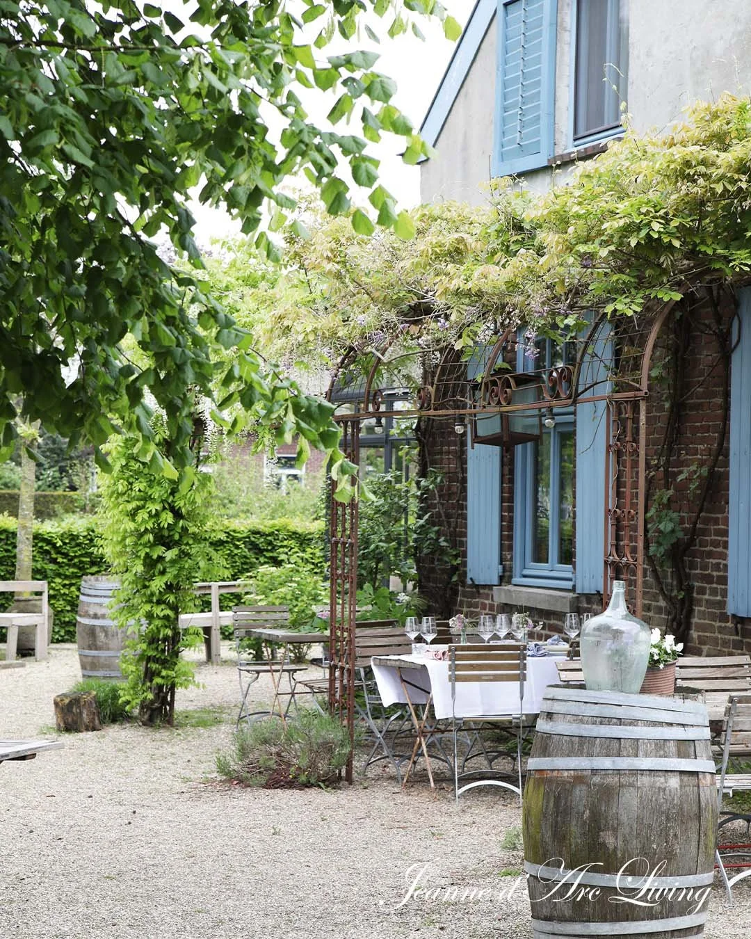 Udendørs café med et bord dækket med en hvid dug, omgivet af træstole, foran en gammel murstensbygning med blå vinduesrammer og blomstrende klatreplanter. Der er grønne træer og buske i baggrunden.