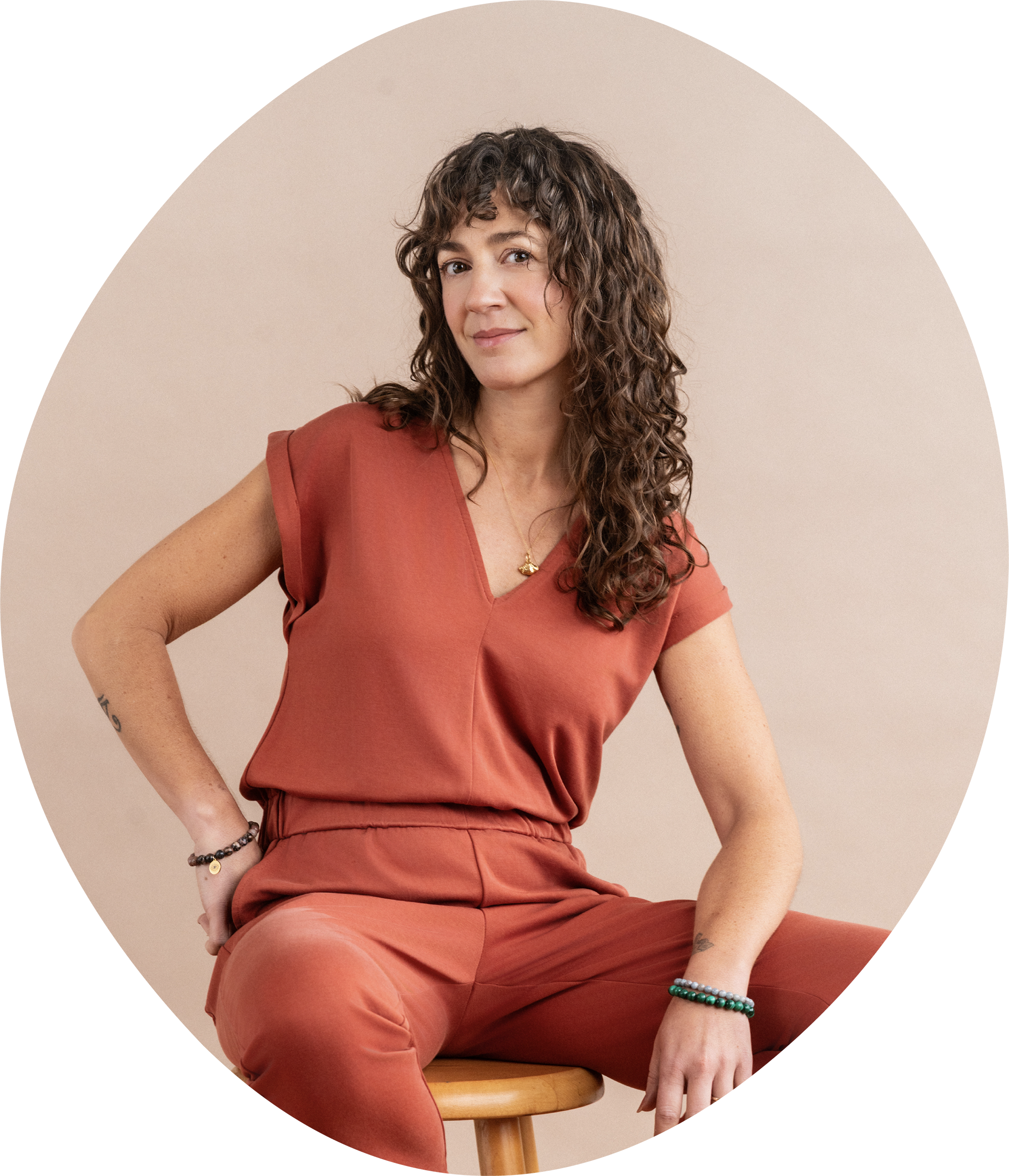 A woman with curly brown hair wearing a rust-colored sleeveless top and matching pants sits on a wooden stool against a beige background, looking confidently at the camera.