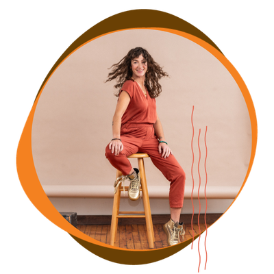 Smiling woman with curly hair sitting on a wooden stool, wearing a rust-colored outfit, in front of a plain beige background.
