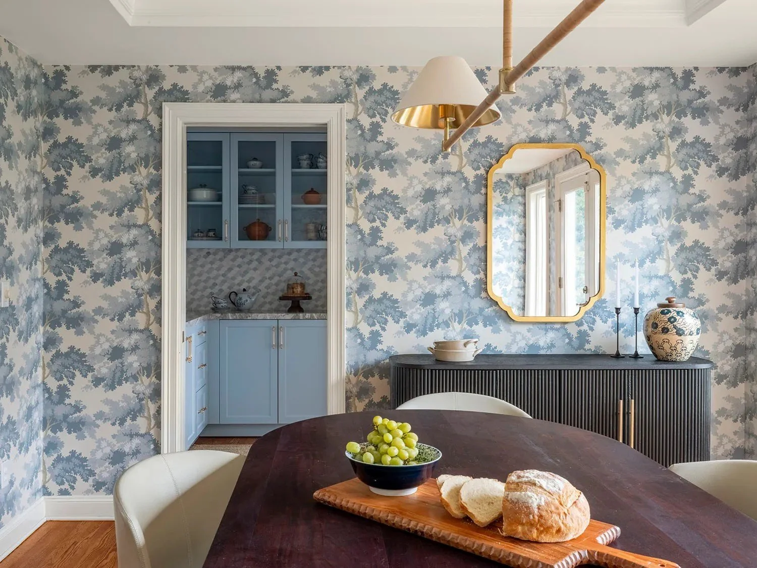 Blue botanical wallcovering wraps the room in pattern and gives the space a softer, more intimate feeling. White trim and crown molding frame the wallpaper and preserve the architectural character of the room, while the dark dining table and fluted s