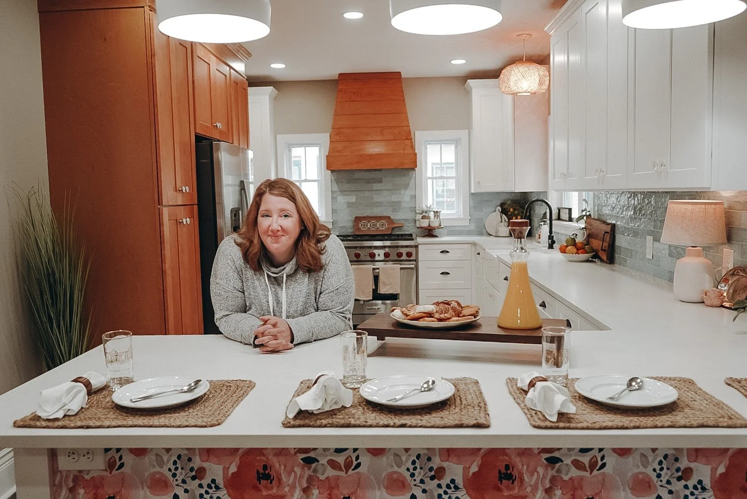 A woman with red hair sitting at a white kitchen island with set plates, napkins, and glasses, in a modern, bright kitchen with wooden and white cabinets, a stove with a wooden hood, and various kitchen decor.