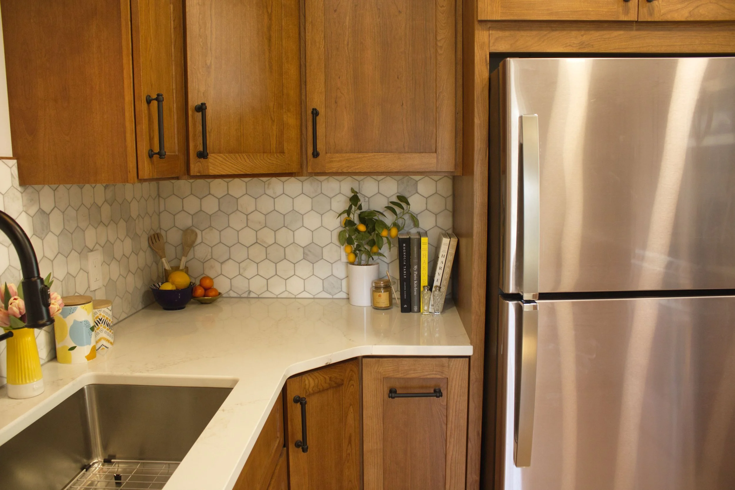 A kitchen with wooden cabinets, a white countertop, and a stainless steel refrigerator. There are a bowl of lemons and oranges, kitchen utensils, and a small bookshelf with books on the counter.