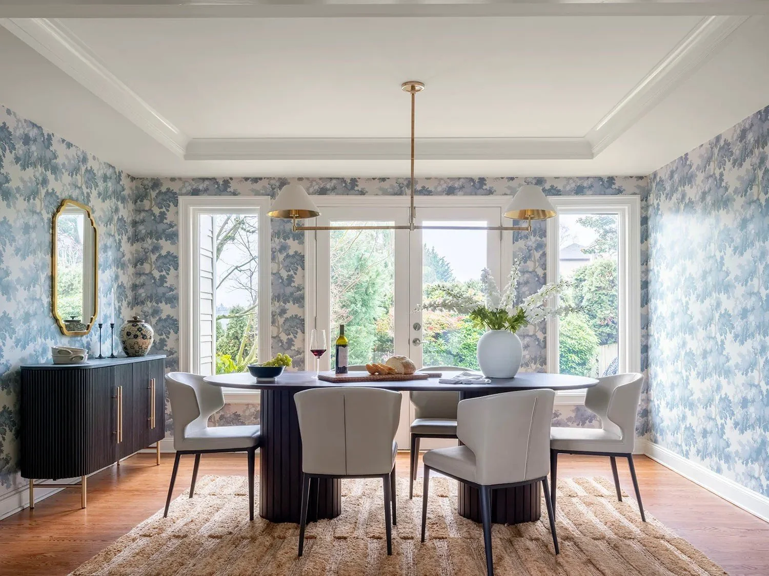 Blue botanical wallcovering wraps the room in pattern and gives the space a softer, more intimate feeling. White trim and crown molding frame the wallpaper and preserve the architectural character of the room, while the dark dining table and fluted s