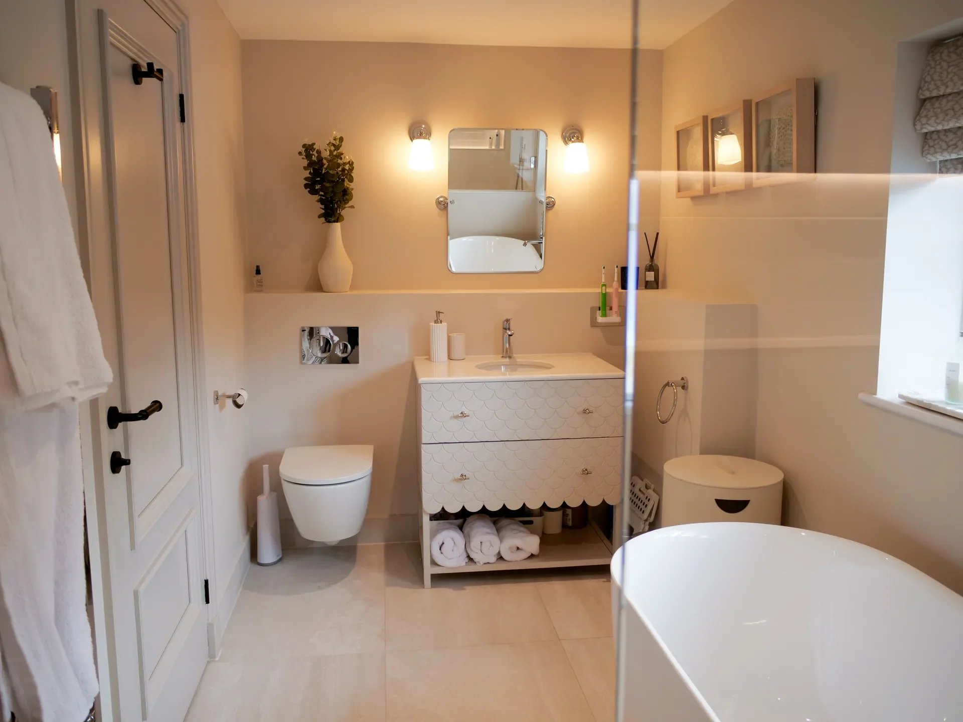 A modern bathroom with a white toilet, a vanity with a mirror, and a shelf with rolled towels. There is a potted plant on the shelf, and the walls are painted in a soft beige color. The bathroom features wall-mounted lights and a window with patterne