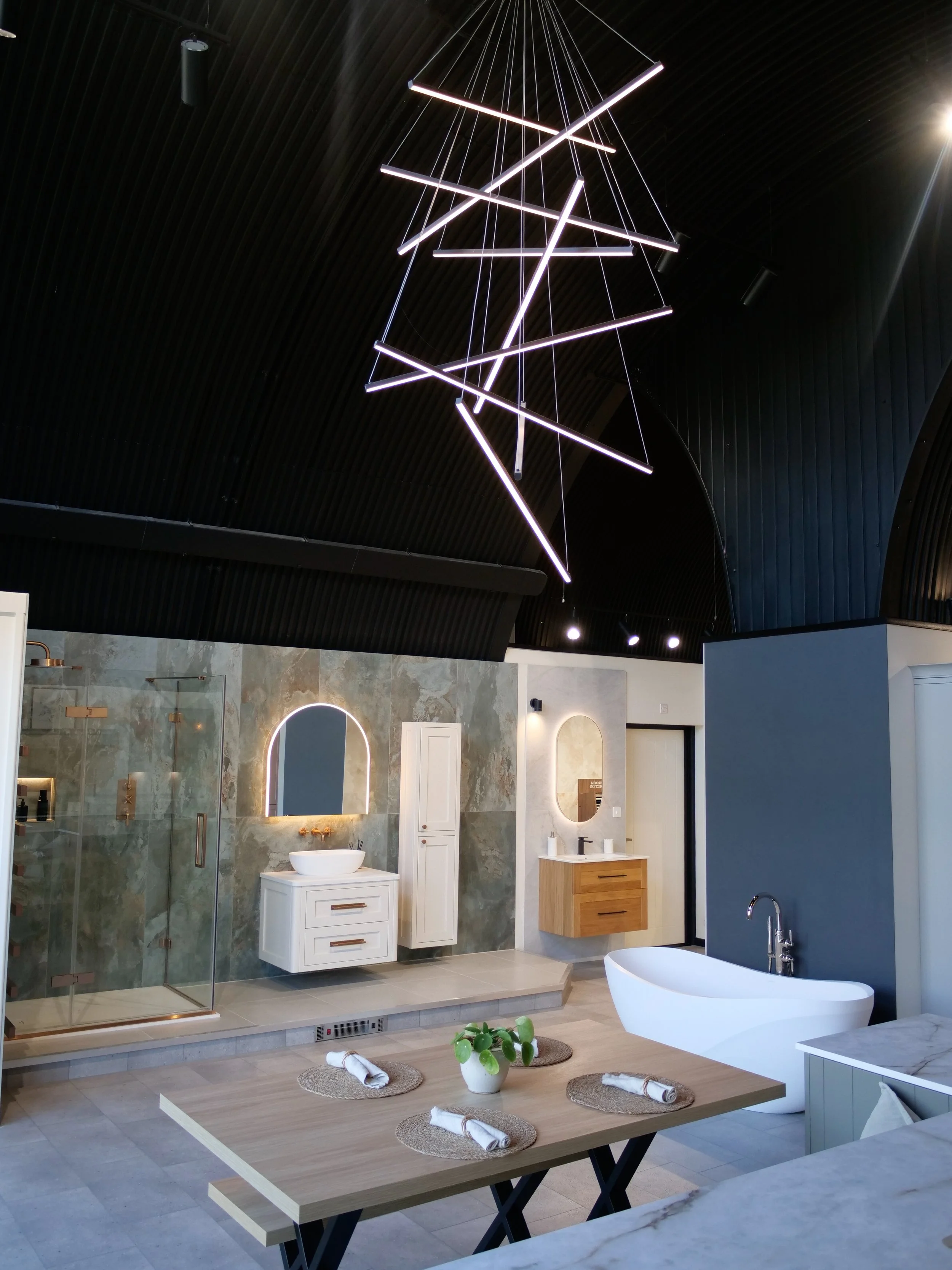 Modern bathroom and dining area with a black ceiling, unique hanging light fixture, floating vanity with a mirror, bathtub, and a wooden table set with placemats and a potted plant.