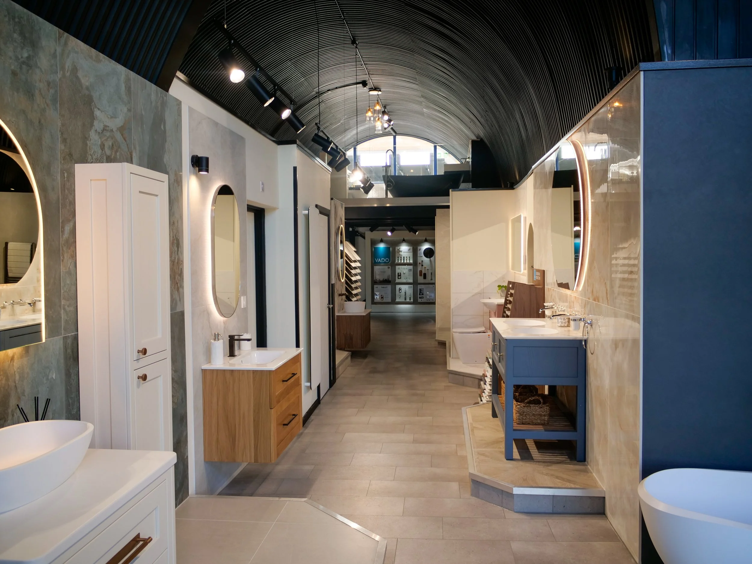 A showroom with various modern bathroom vanities, sinks, and mirrors displayed in a brightly lit space with a curved black ceiling.