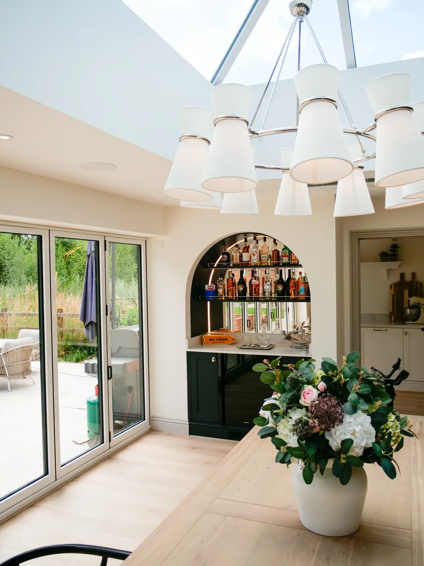 Living room with large sliding glass doors leading to an outdoor patio, a wall-mounted bar shelf with various liquor bottles, a modern chandelier overhead, a large flower arrangement in a white vase, and a glimpse of the kitchen in the background.
