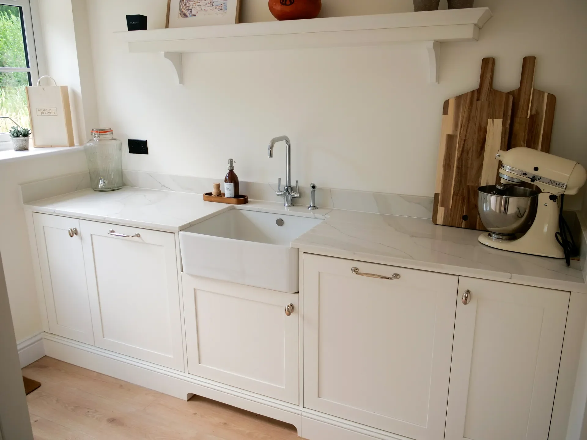 White kitchen cabinet with a marble countertop, sink, and faucet. On the countertop, there are a soap dispenser and a small tray. Behind the sink, there are two decorative cutting boards leaning against the wall. To the right, there is a beige stand 