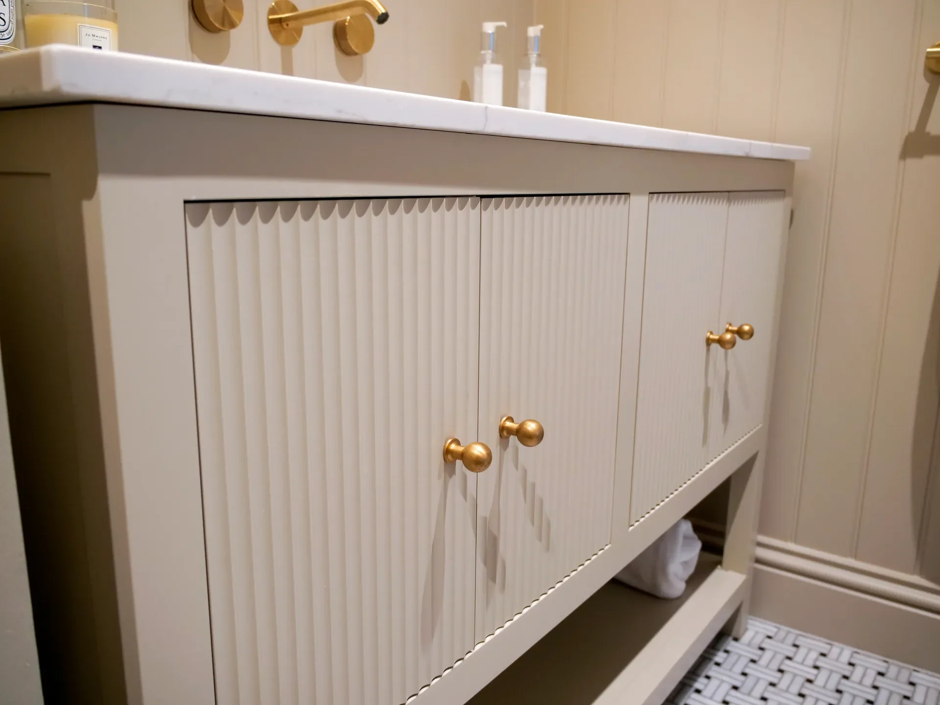 A white bathroom vanity with slatted cabinet doors and gold round knobs, with a marble countertop and gold fixtures, located in a bathroom with beige walls and a white tile floor. Two lotion bottles are visible on the countertop.