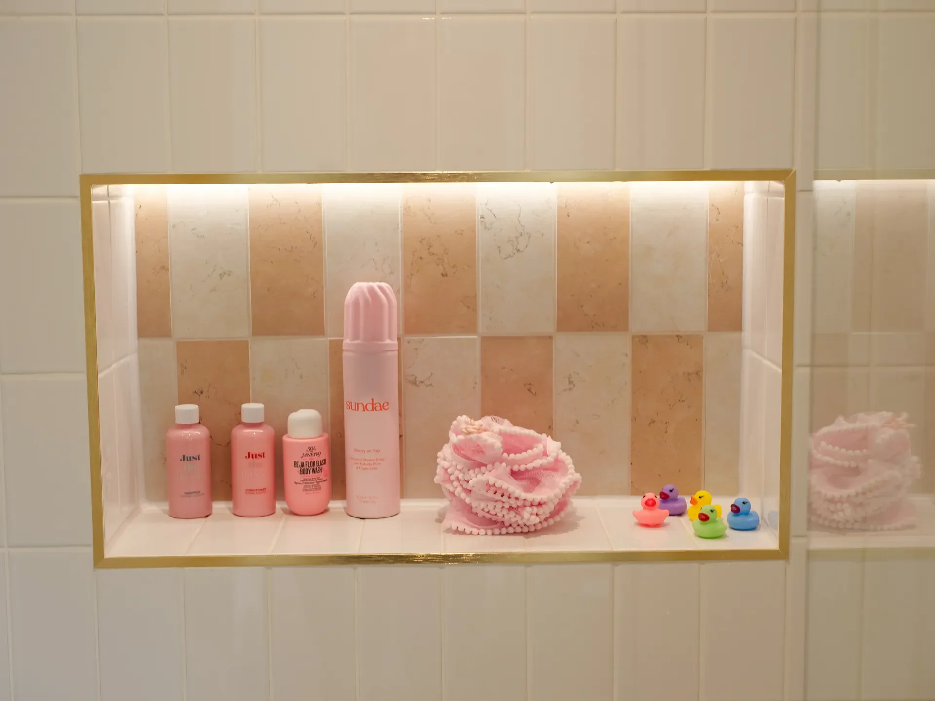 Bathroom shelf with pink bottles labeled 'Just', pink loofah, and small colorful rubber duck toys.