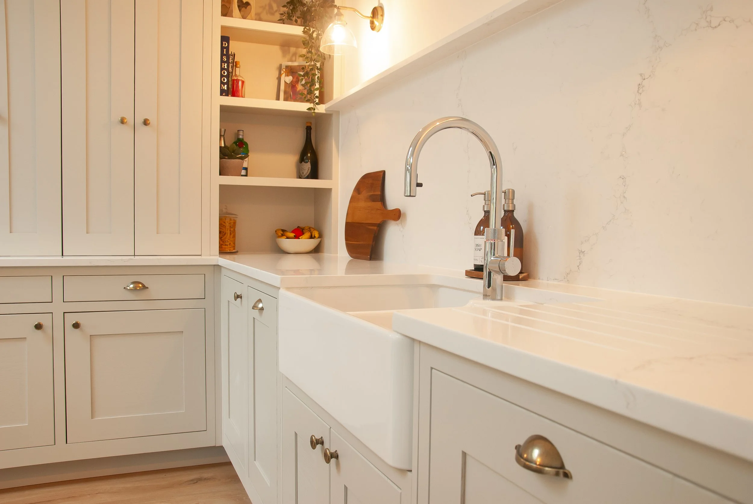 A kitchen with white cabinets, a farmhouse sink, a modern chrome faucet, open shelves with decorative items and bottles, and a marble countertop.