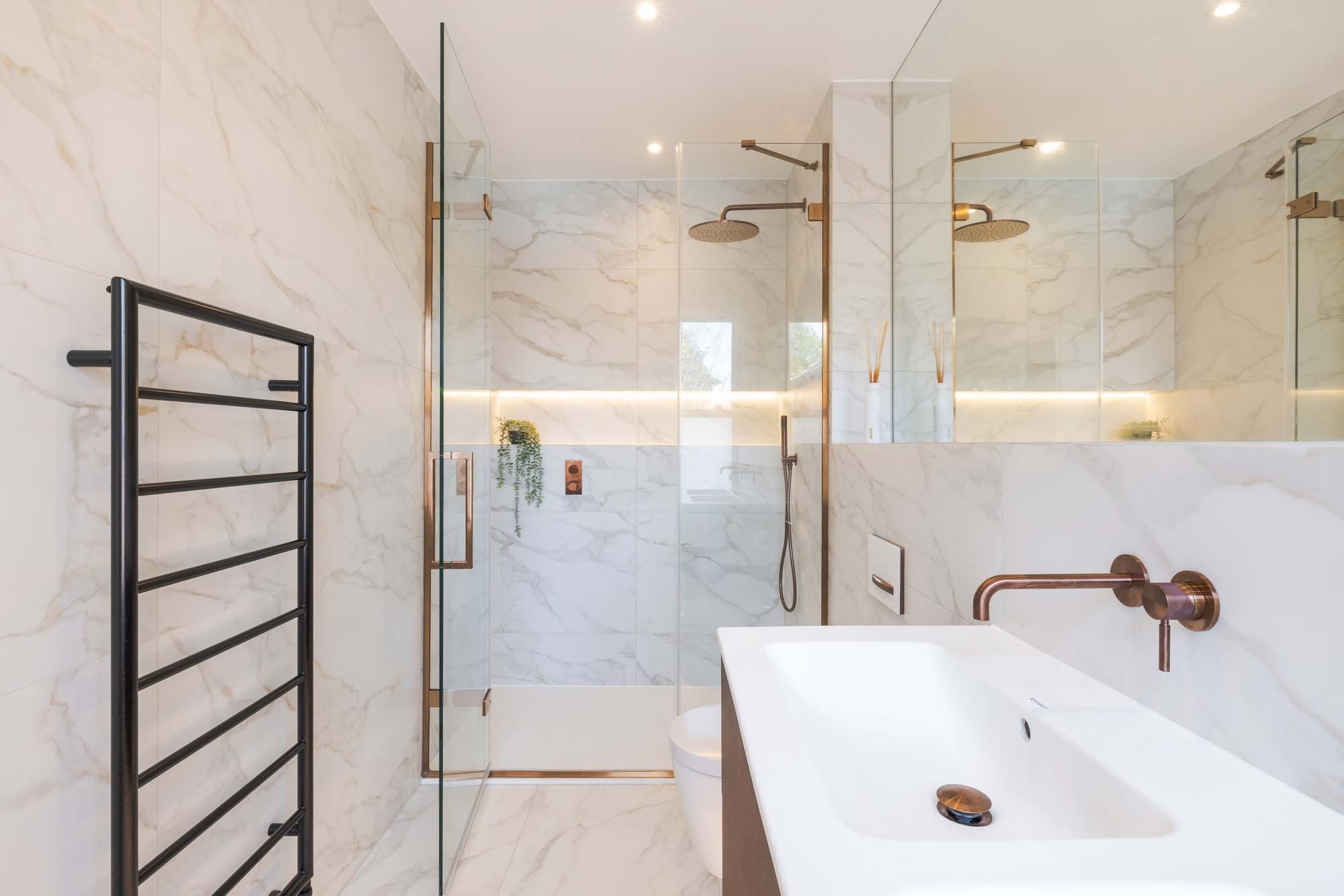 Modern bathroom with marble walls and floor, glass shower enclosure, black towel rack, white sink with copper faucet, large mirror, and bronze shower fixtures.