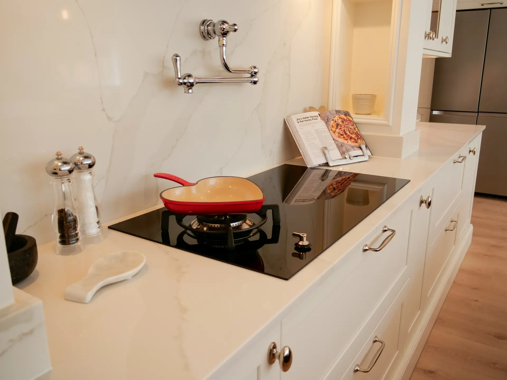 Kitchen countertop with a red frying pan on a black stove, salt and pepper shakers, a napkin, an open cookbook with a pizza recipe, and a breakfast bowl on a windowsill, with white cabinets and stainless steel appliances.