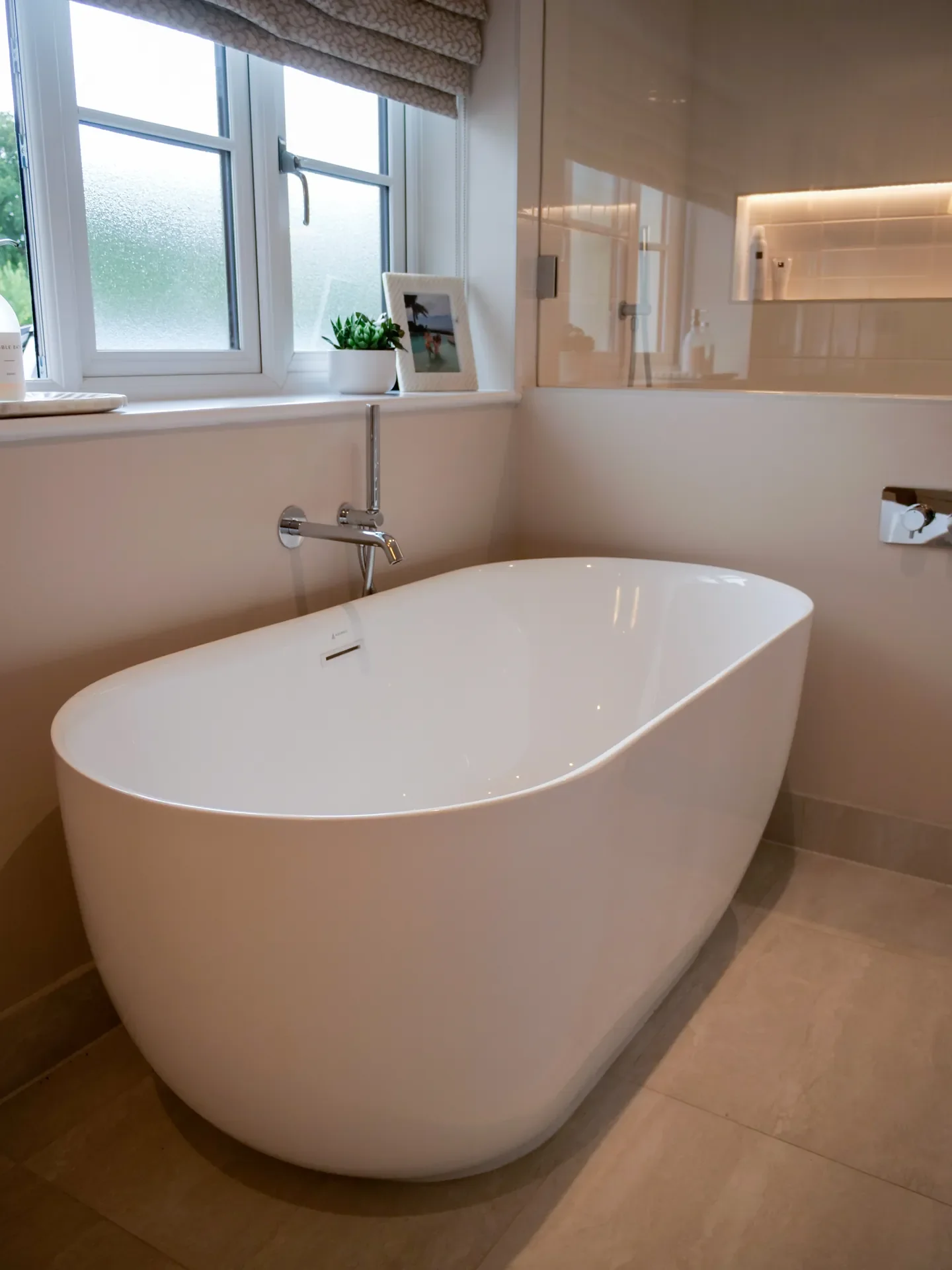 Modern bathroom with a freestanding white bathtub, a window with frosted glass, a small potted plant, and a framed photo on the windowsill.