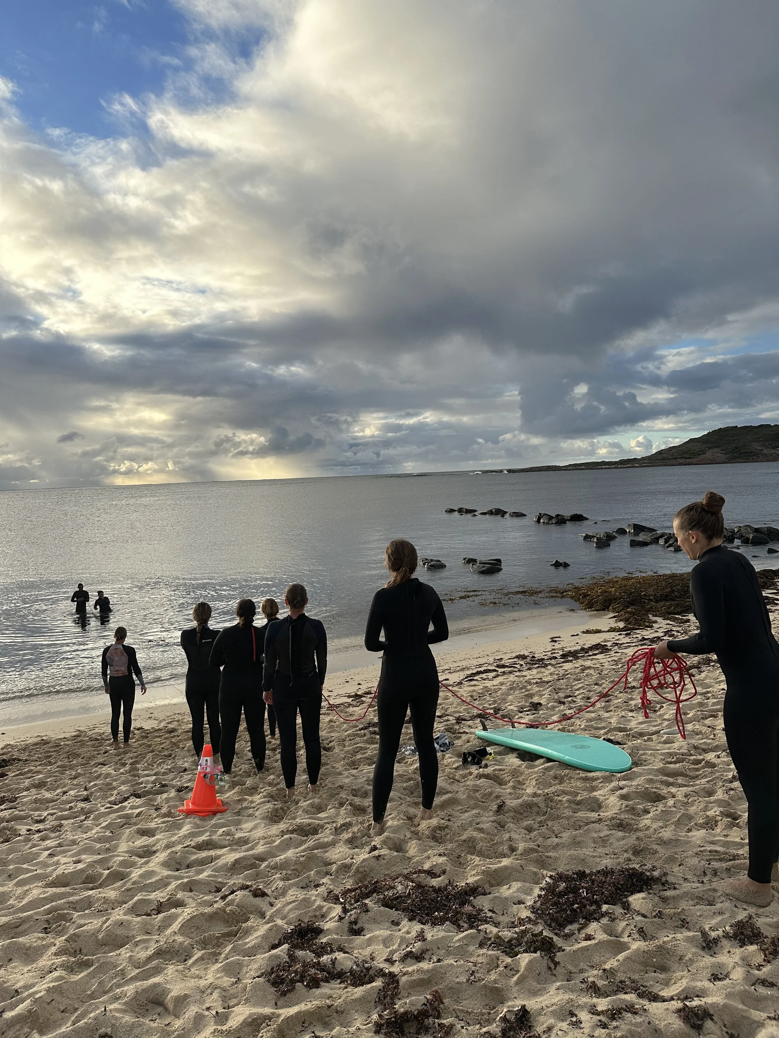 Group of people in wetsuits on a beach preparing for a surf session, with some in the water and surfboards on the sand under a cloudy sky.