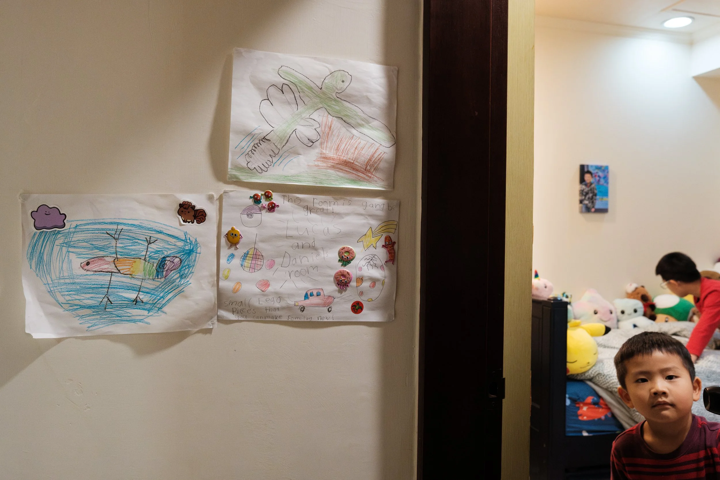A little boy is looking through the doorway of his bedroom in Linkou, Taiwan. There are hand-drawn pictures and signs on the wall next to the door.