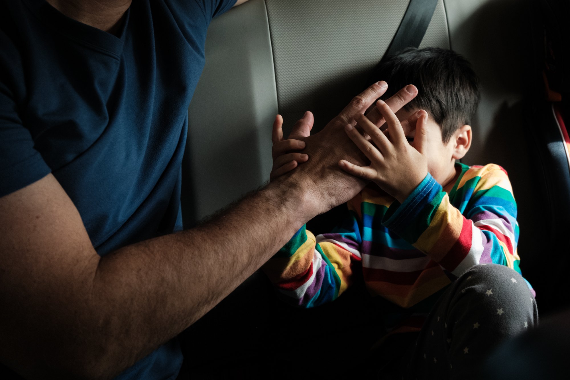 A dad plays with his son in the backseat of a car and covers his face with his hand during a road trip in Taipei, Taiwan.