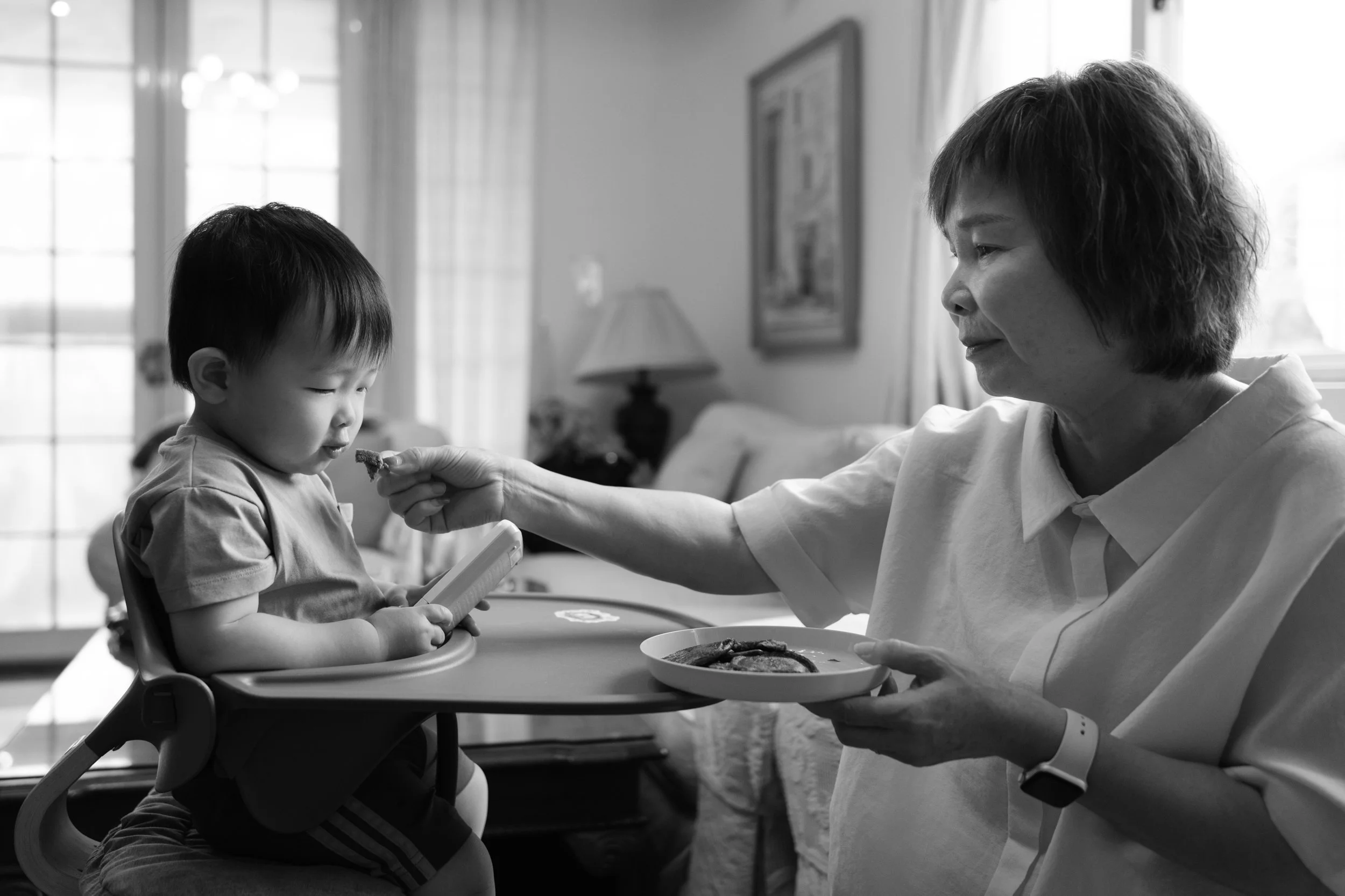 A black and white image of a grandmother feeding her toddler grandson. The boy is sitting in a high chair with light shining on him from a window.