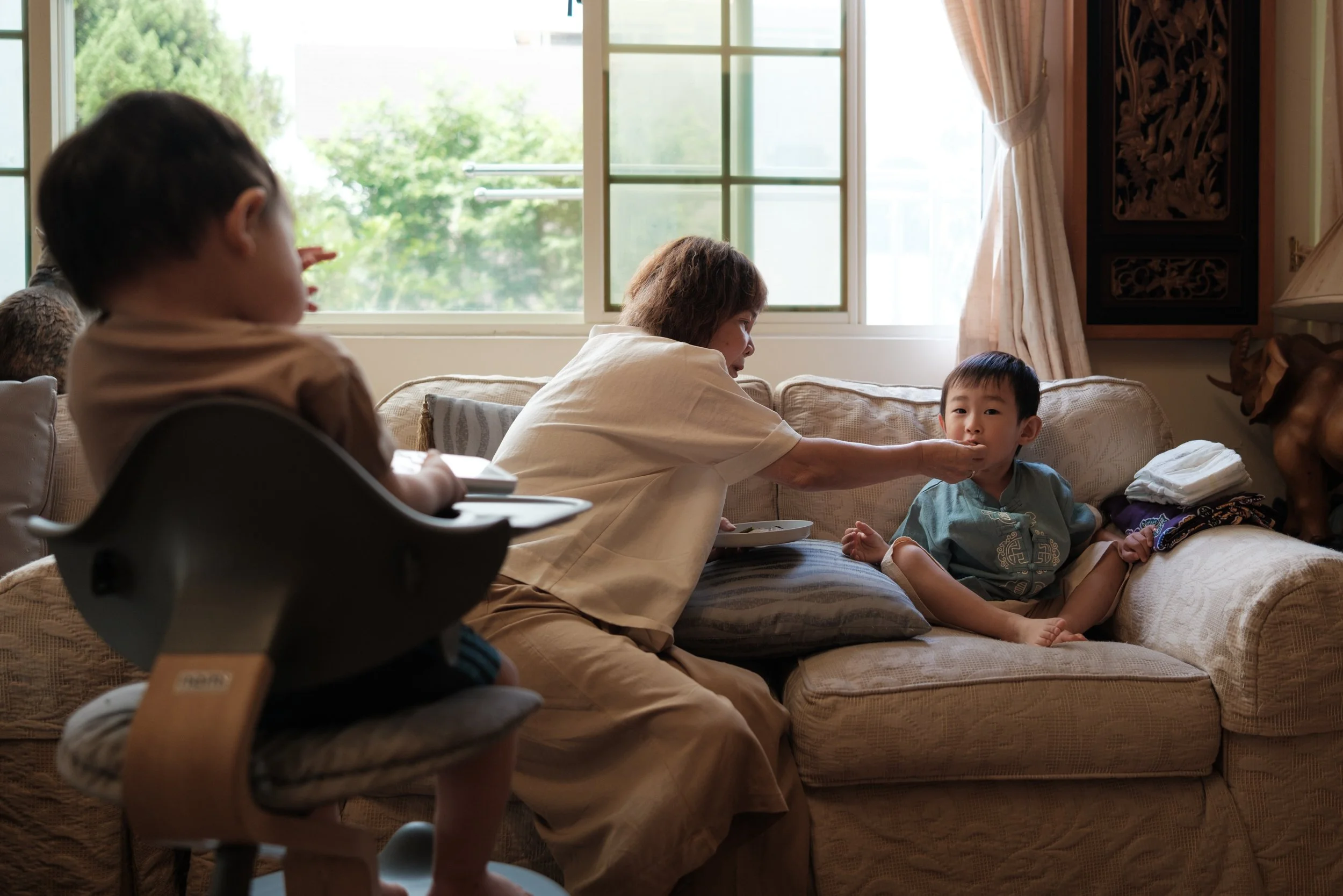 A color image of a grandmother feeding her grandchildren in front of a window in her living room in Taipei. Captured by family photographer Cahleen Hudson.