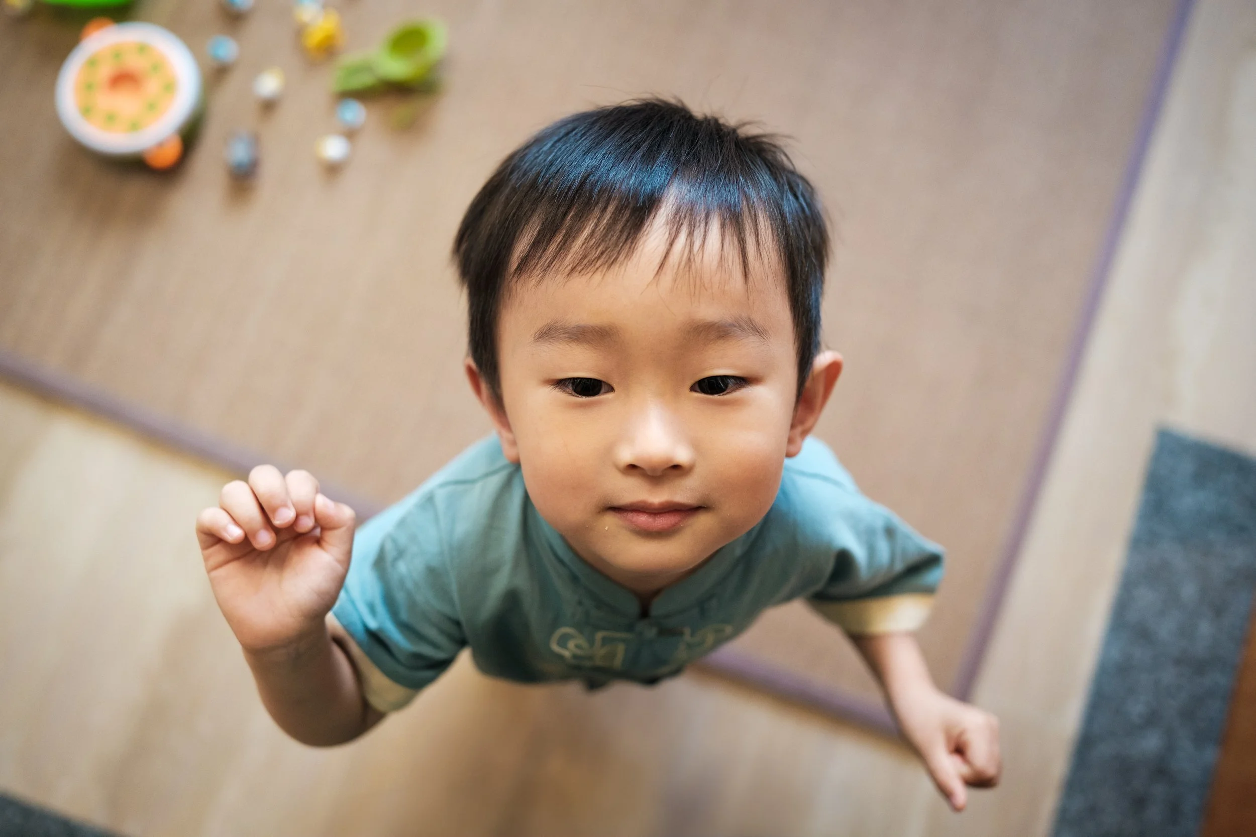 A color photograph of a little boy looking up. There is a tatami mat with toys on it at his feet. Photographed by Cahleen Hudson during a home family photoshoot in Taipei.