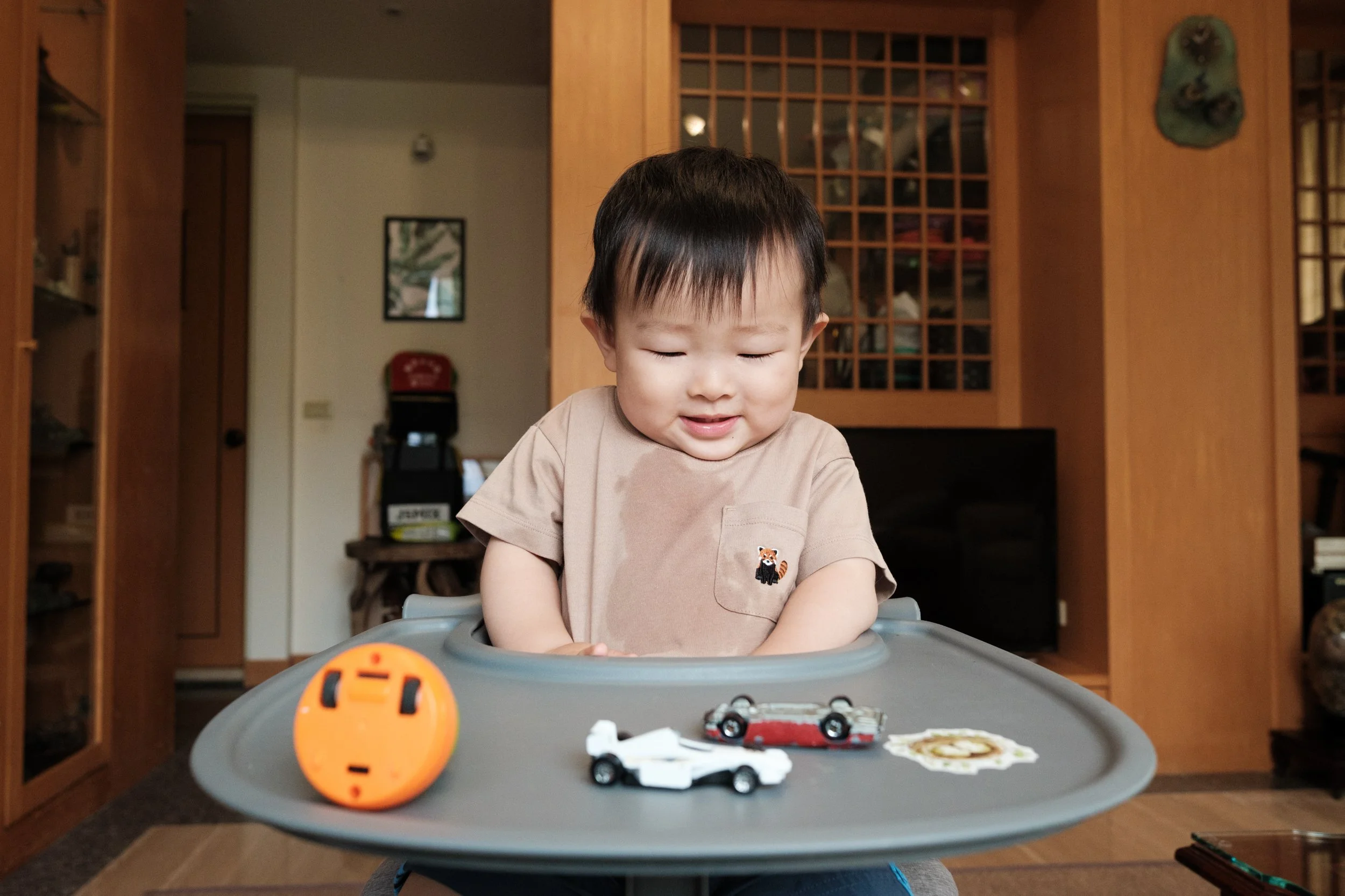 A color photo of a toddler boy in his high chair. He has drool running down his shirt and there are toys scattered across the tray of his high chair.
