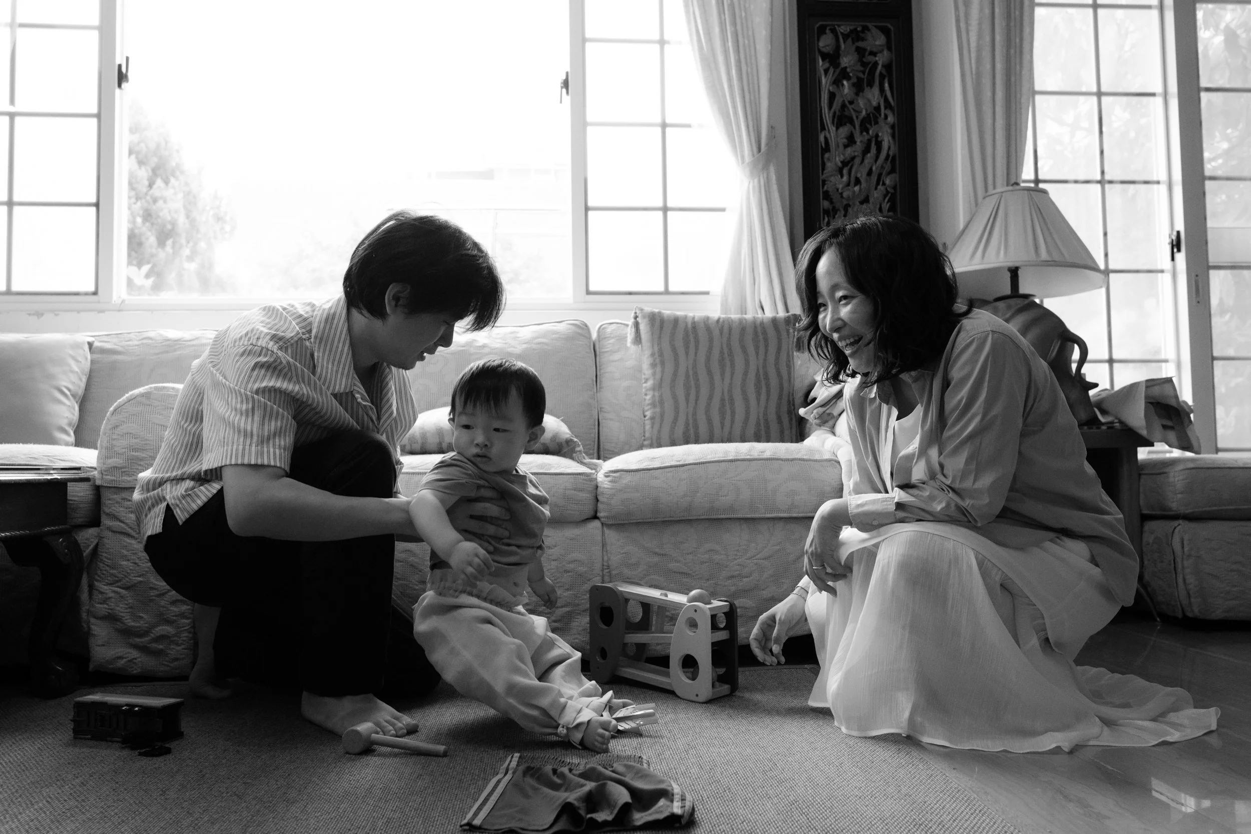 A black and white photo of a couple playing with their toddler son in their living room in Taipei. There is a big window with light streaming in behind them.
