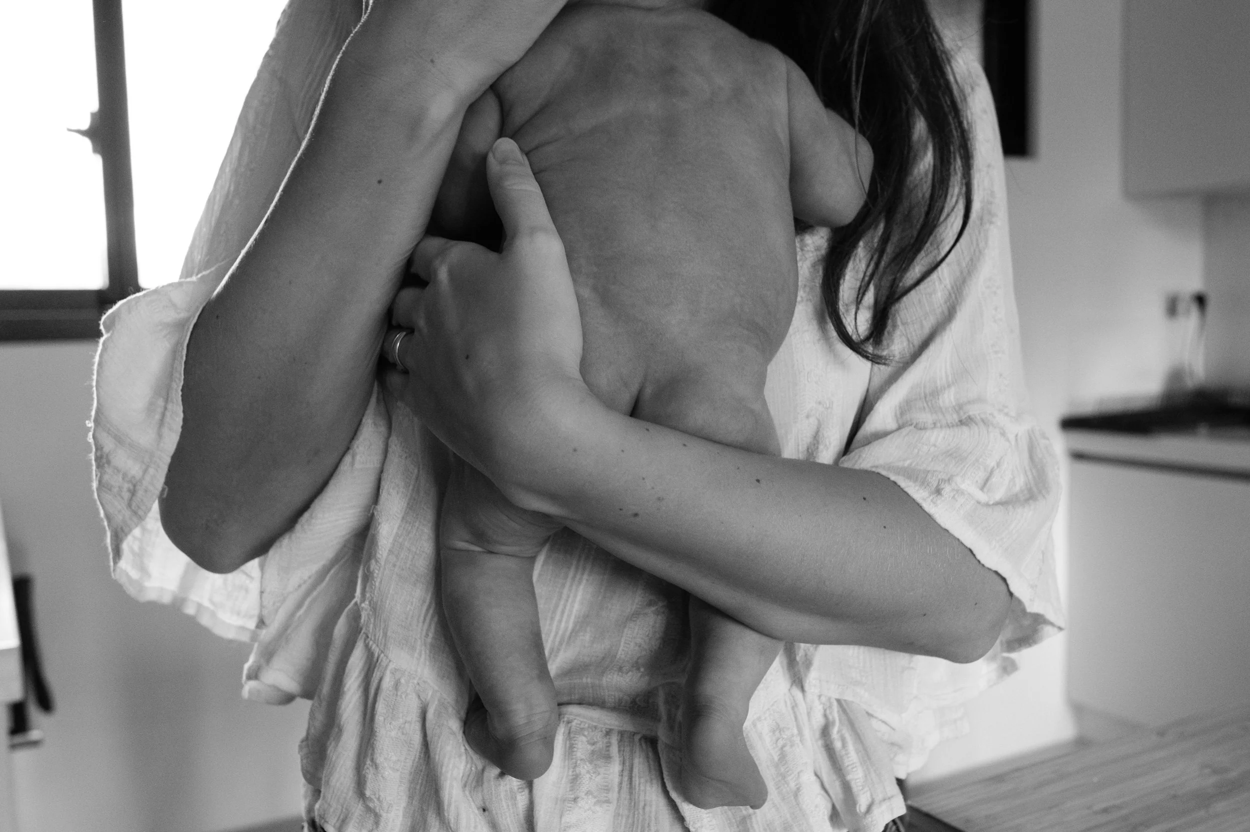 A black and white image of a newborn baby's backside. The baby is being held by his mother, and both of her arms are wrapped around him.