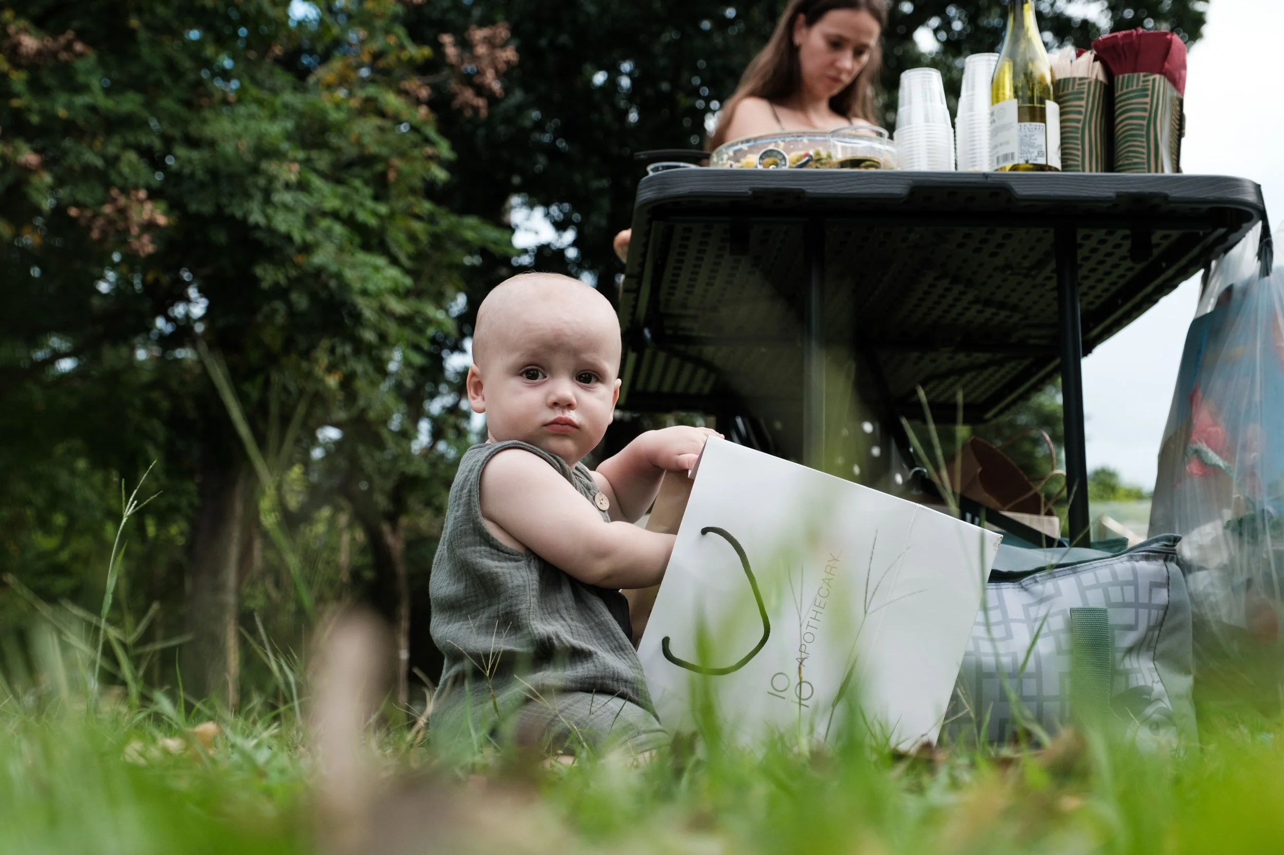 A color photo of a baby reaching into a gift bag at his birthday party. The baby is partially obscurred by tall grass.