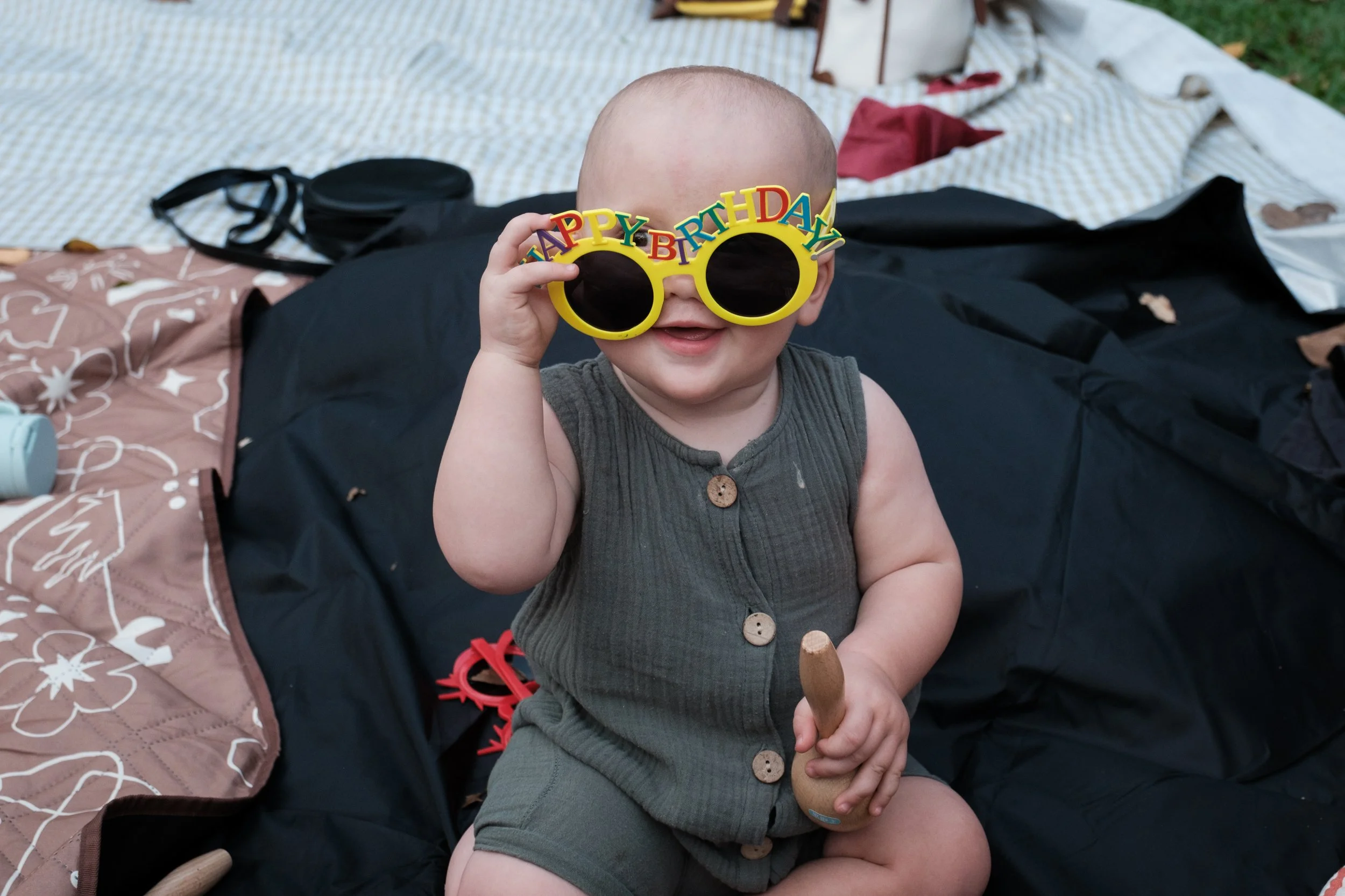 A baby is sitting on a blanket at Daan Park in Taipei. He is peeking over the top of sunglasses that say happy birthday.
