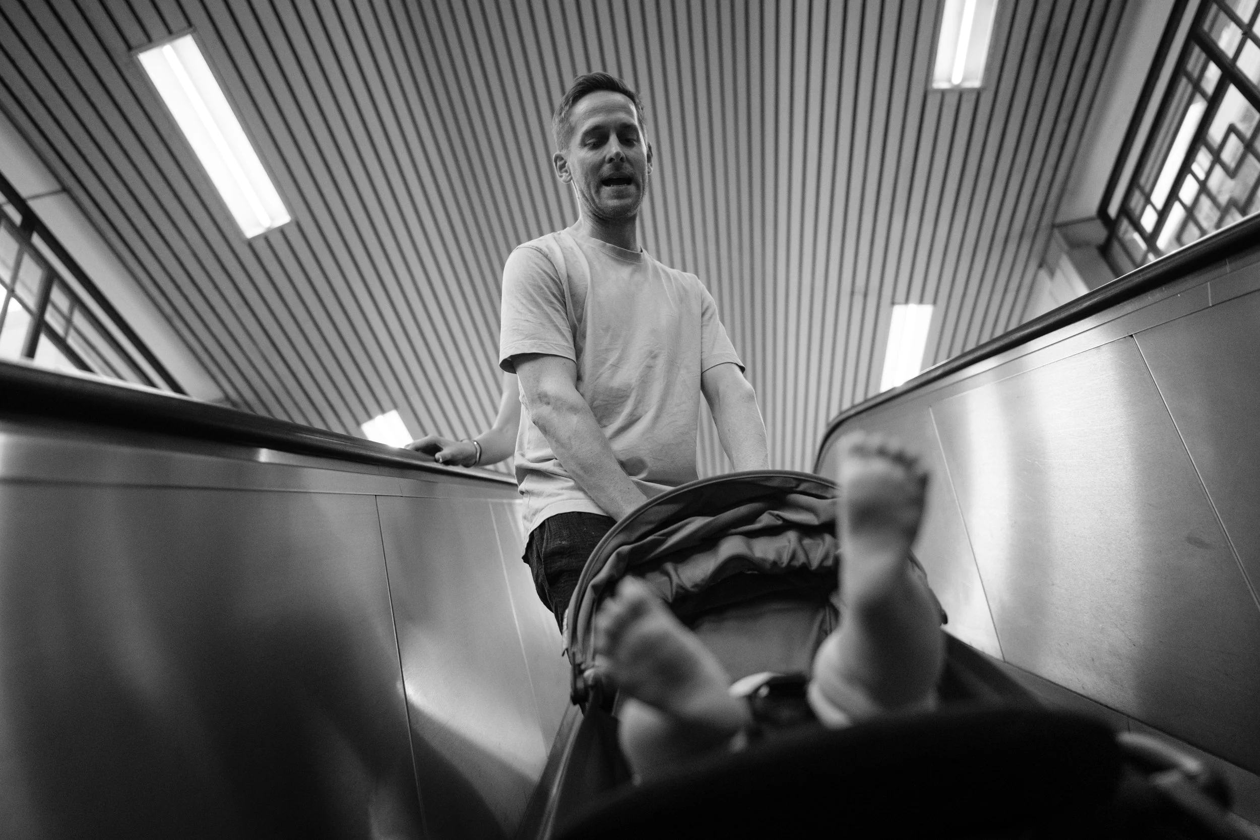 A black and white image of a dad holding onto a stroller as they go down an escalator in a Taipei MRT station. The baby's feet are sticking out of the bottom of the stroller.