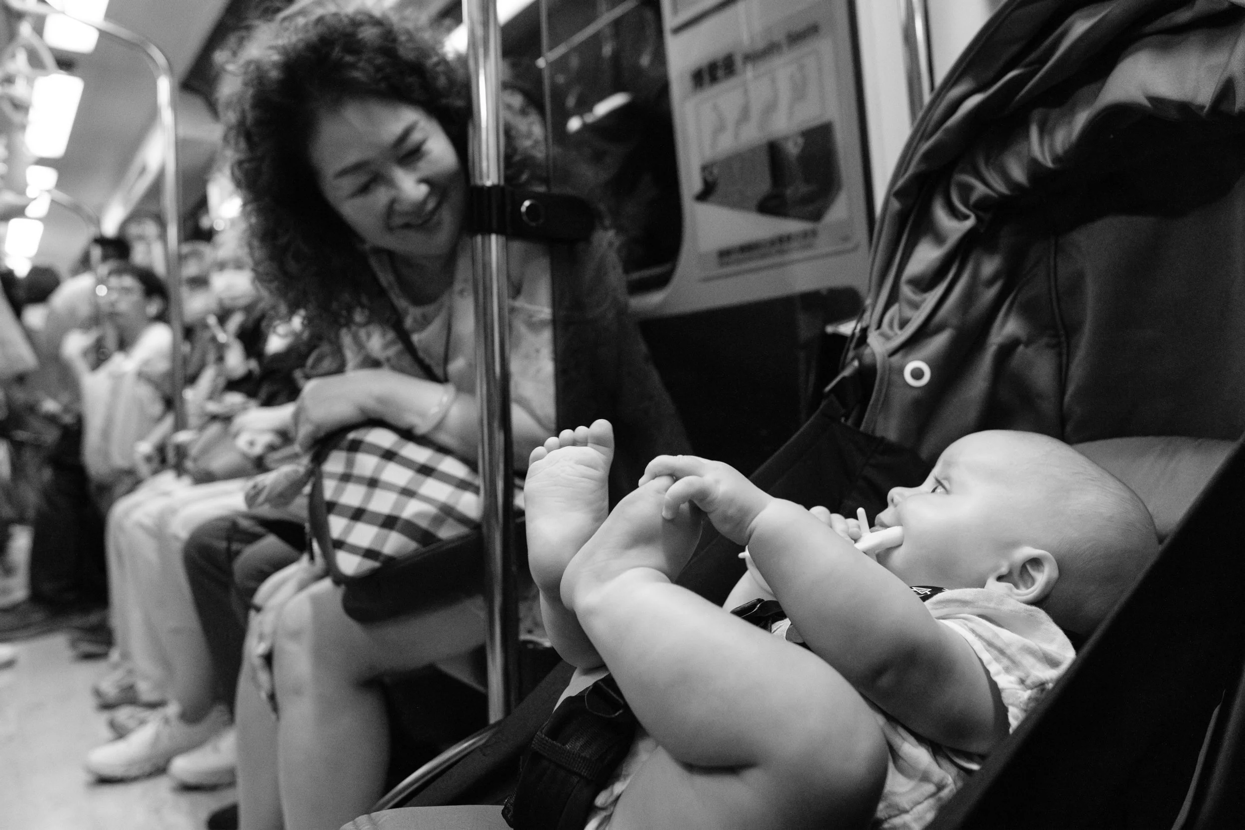 A Taiwanese woman on the MRT in Taipei is looking at the German baby boy in his stroller as he plays with his feet. She is smiling at the baby.
