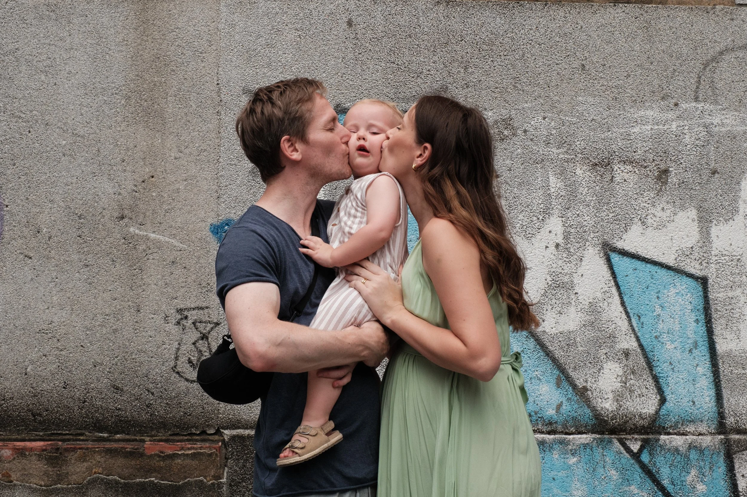 A color image of a mother and father each kissing one of ther son's cheeks as they hold him. The boy's cheeks are being squished and you can see a wall with graffiti behind them.