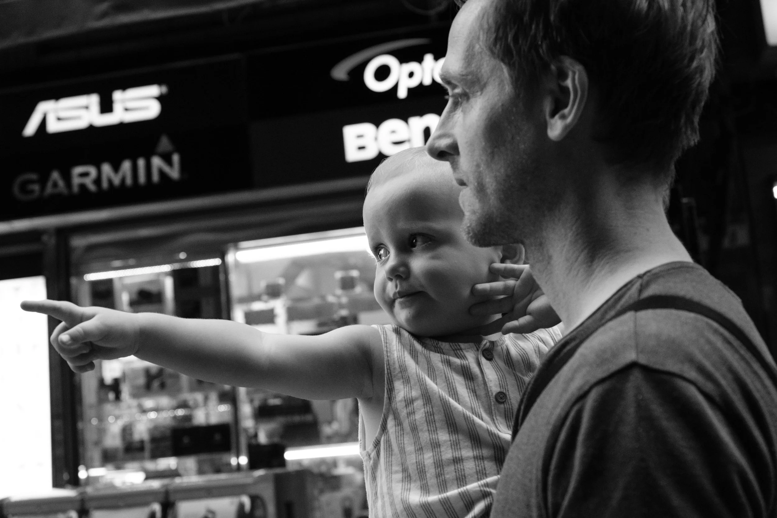 A black and white image of a father holding his toddler son as they walk down a Taipei street. The boy is pointing at something in front of him. Photographed by family photographer Cahleen Hudson.