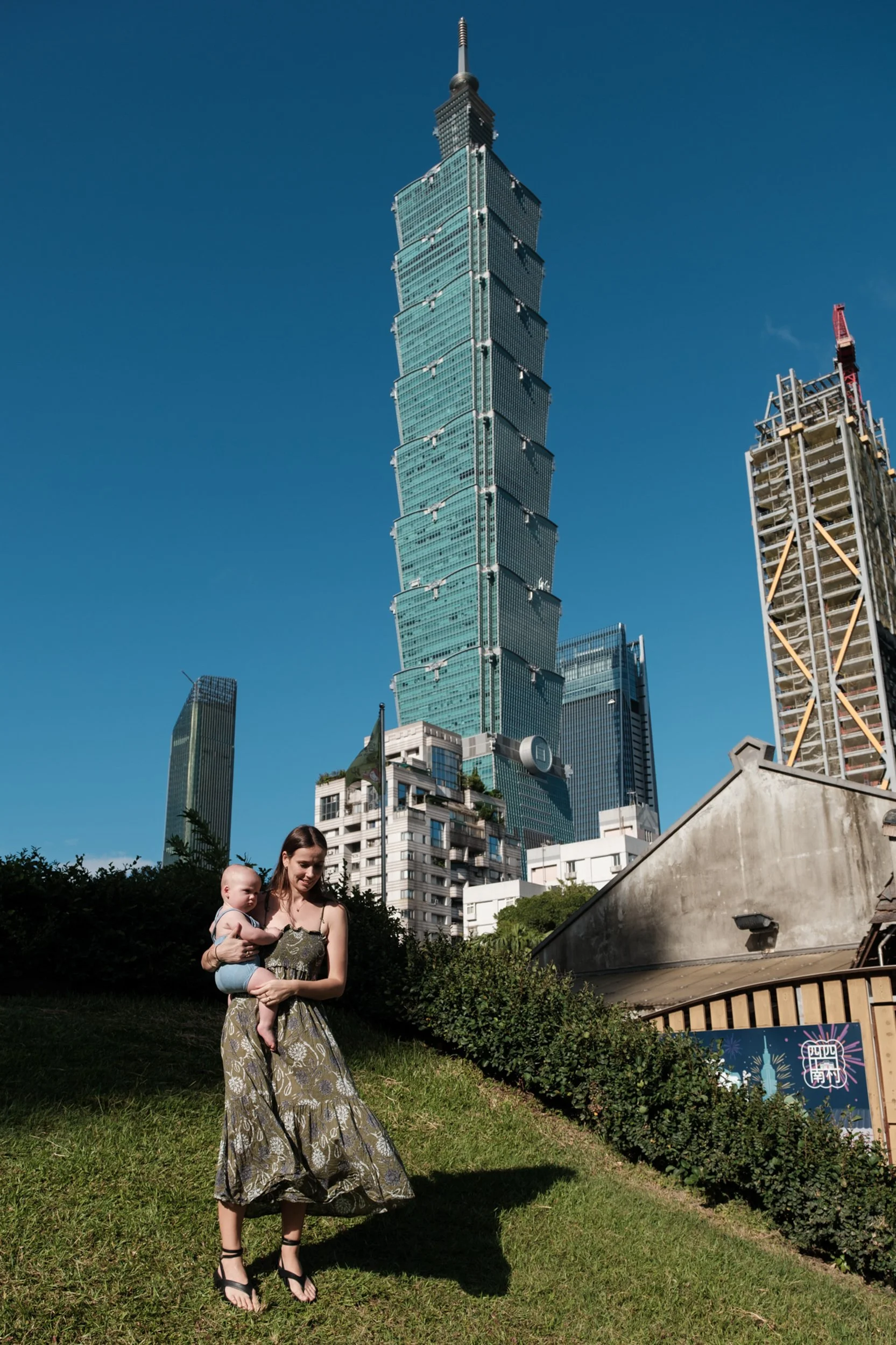 I woman is holding her baby son on a grassy hill with Taipei 101 in the background. Photographed by Taiwan family photographer Cahleen Hudson.
