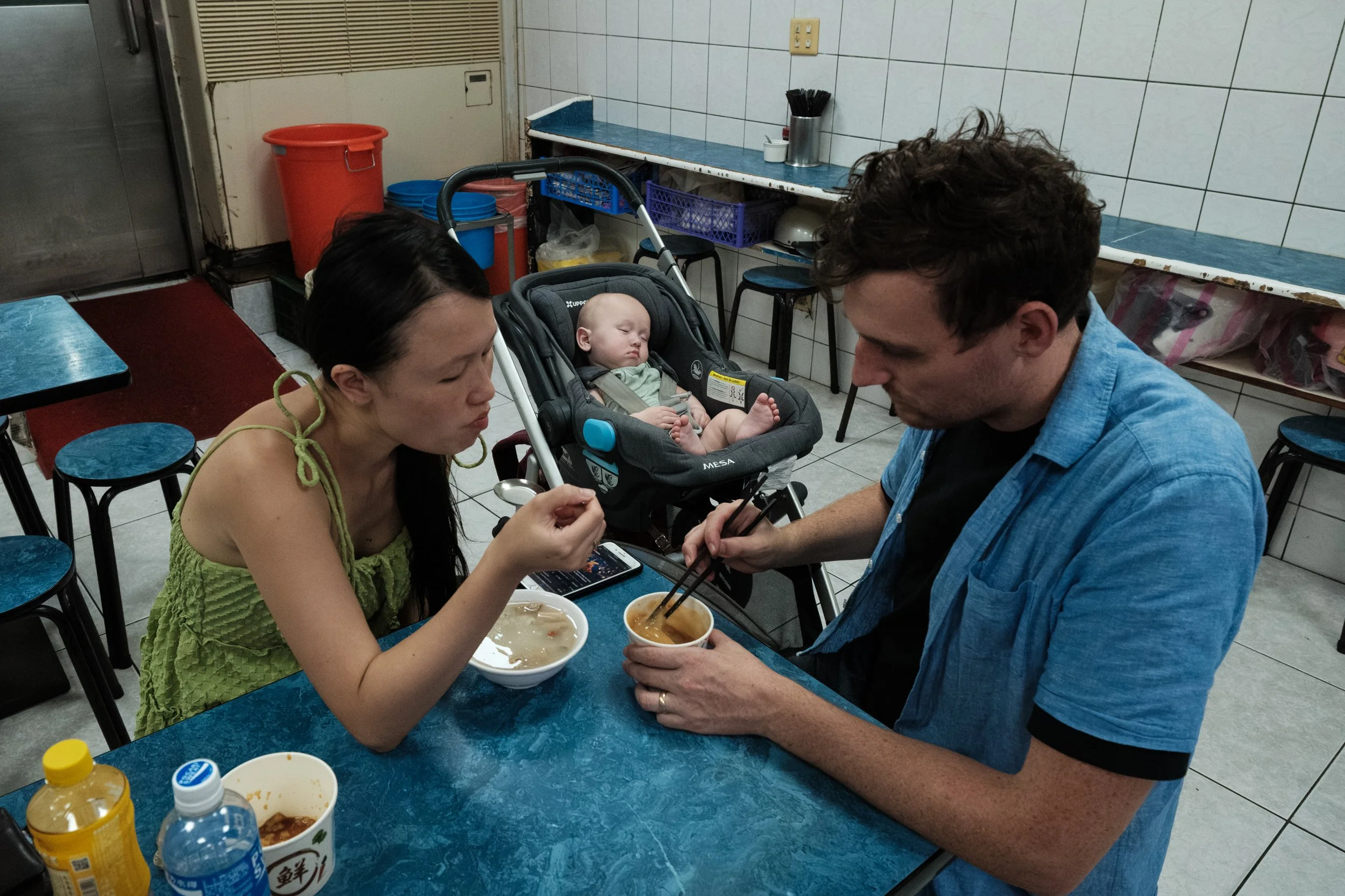 A couple eating noodles at a table in a small Taipei restaurant. Their baby can be seen sleeping in a stroller in the background.