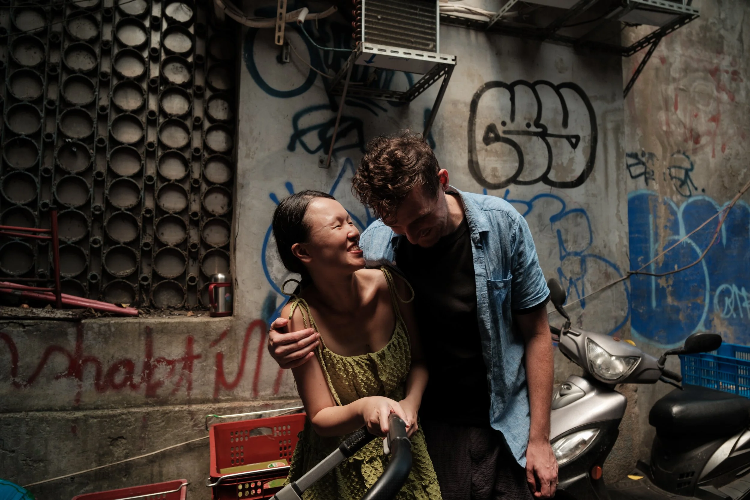 A couple is laughing with a graffiti-covered wall in the background. The man's arm is around the woman's shoulder. 